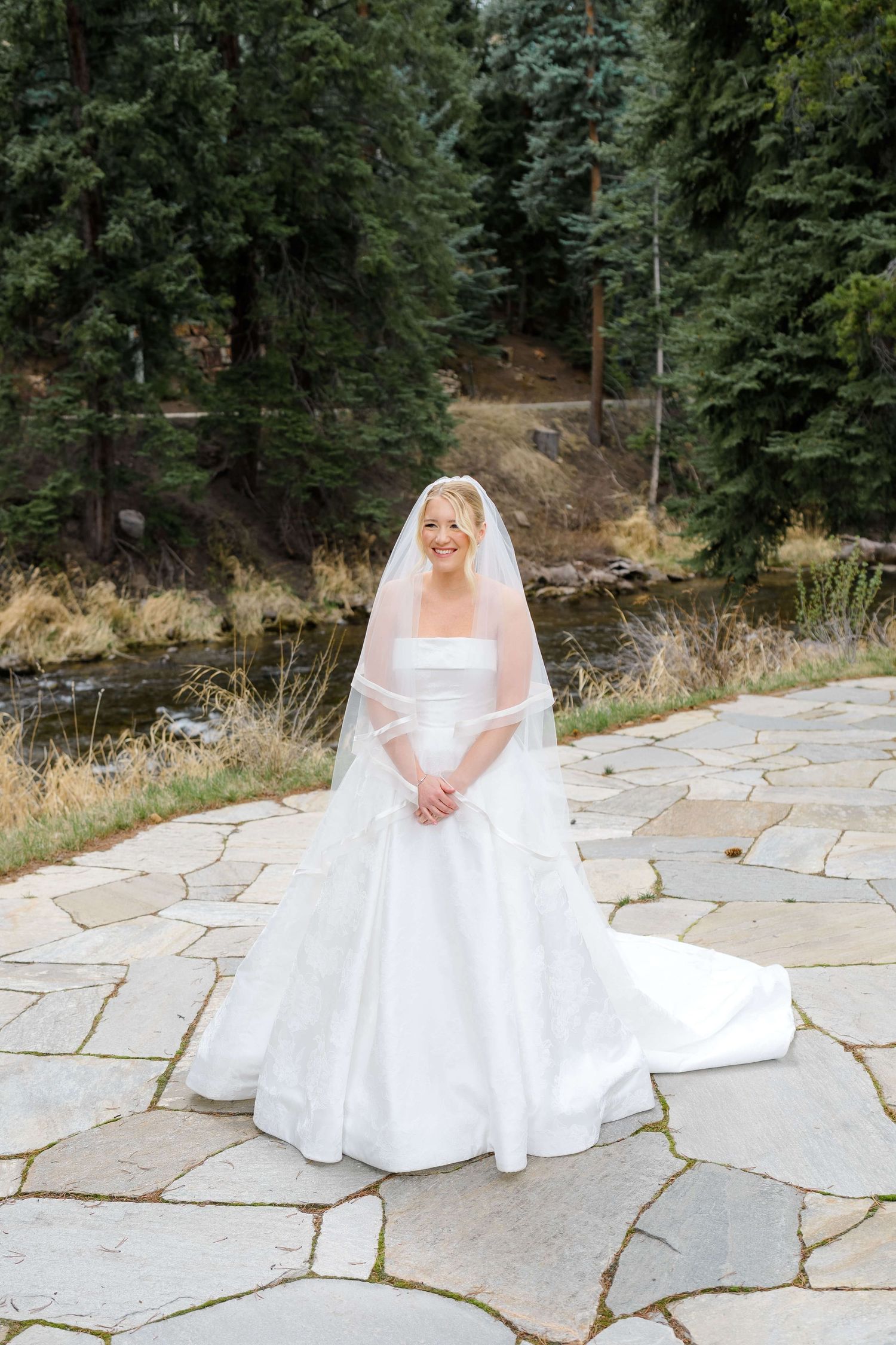 Bride in white ball gown and veil stands on stone patio surrounded by evergreen trees near a stream at Vail Sonnenalp Wedding.