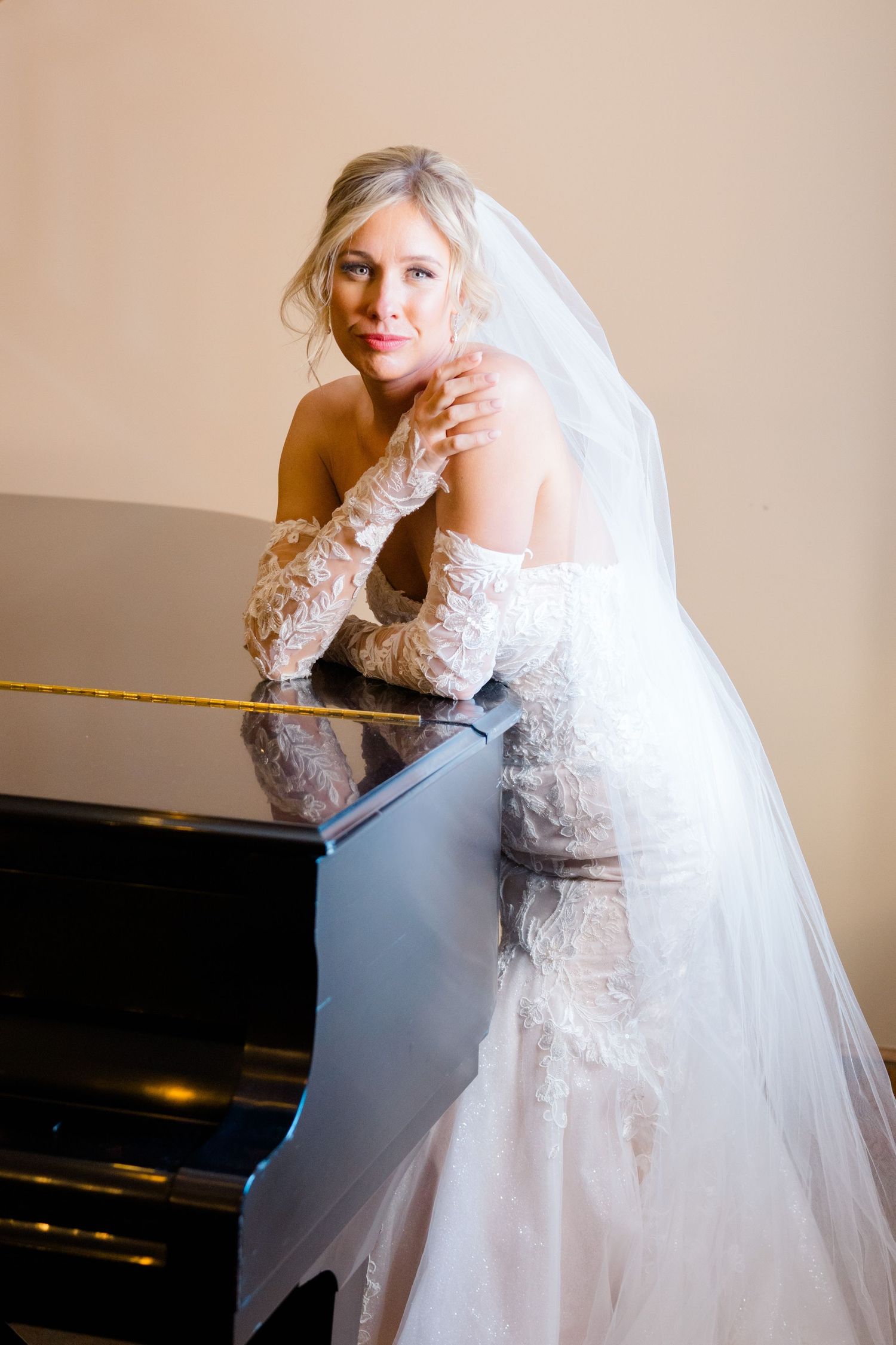 A bride in an elegant lace wedding gown with long sleeves and flowing veil poses gracefully by a piano in vail wedding.