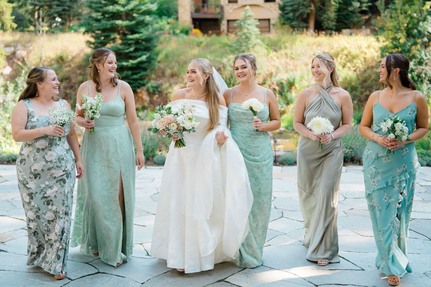 Bride in white gown walks with five bridesmaids in sage green dresses, laughing outdoors at a garden wedding.