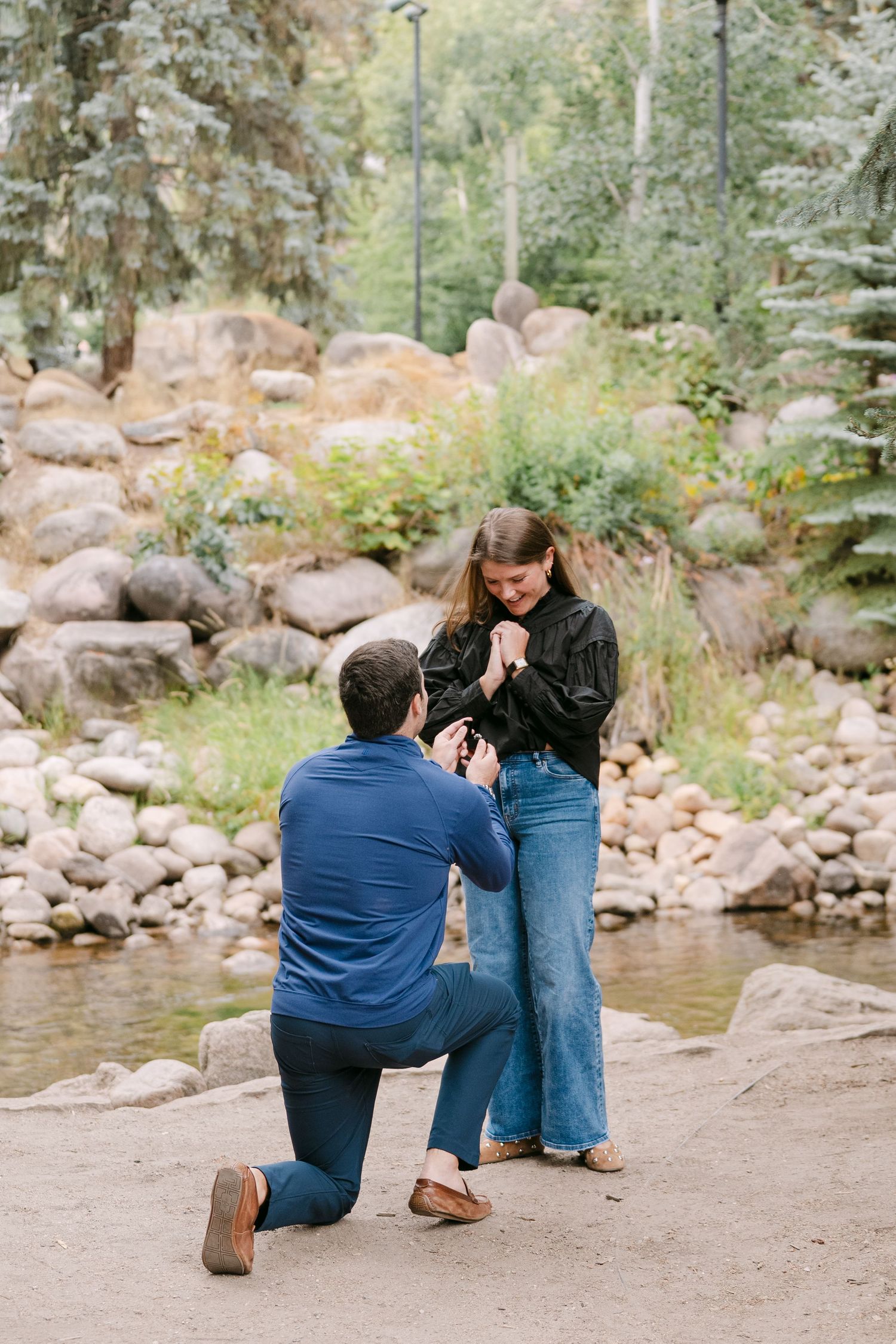 A romantic proposal sequence by a serene mountain lake with rocky shoreline and evergreen trees in the background.