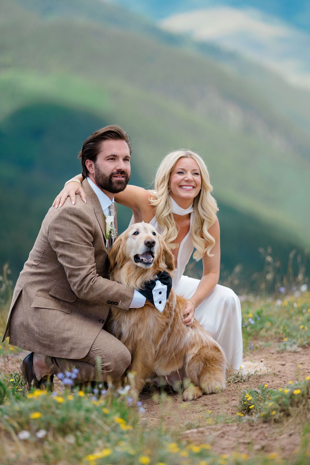Bride and groom pose with golden retriever wearing tuxedo collar on mountain meadow wedding day.