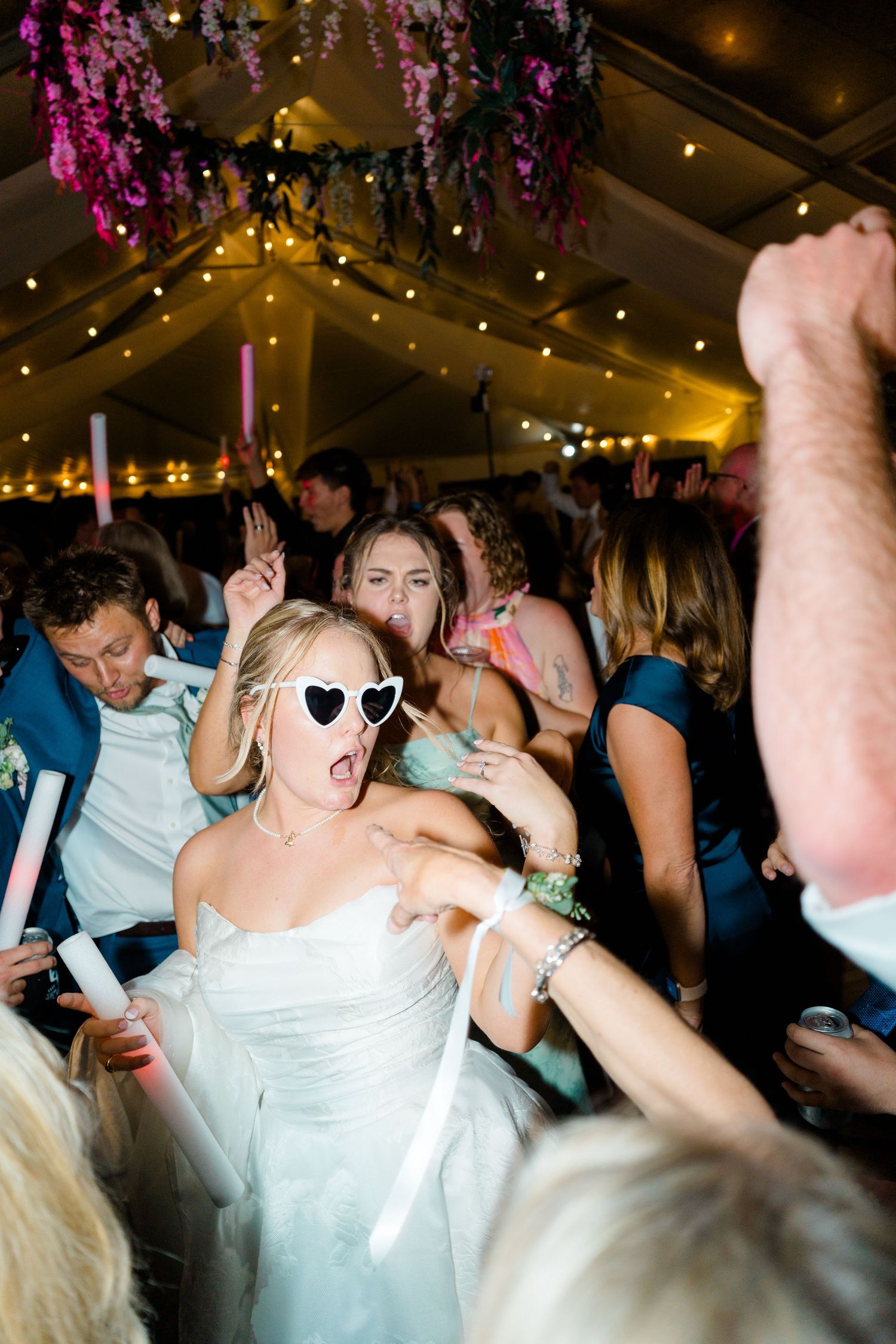Wedding reception dance party with guests dancing energetically under string lights and purple floral decorations.
