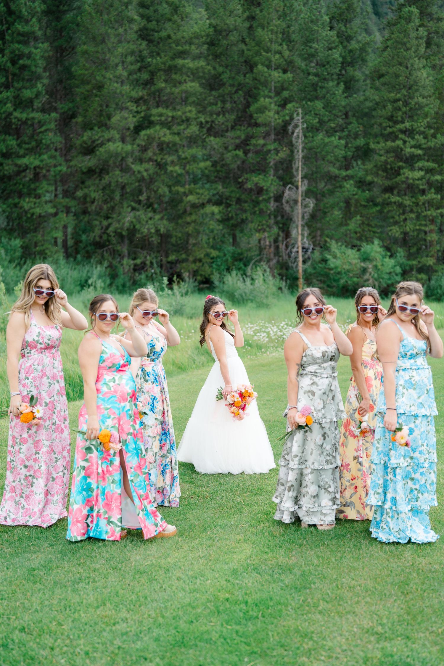 Bridesmaids in floral print dresses gather on grass with mountain scenery and evergreen trees behind them.