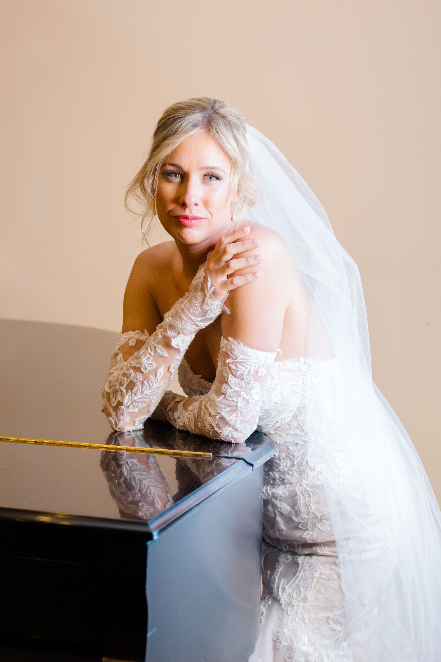 A bride in a white lace wedding gown with long gloves leans elegantly against a piano in a romantic bridal portrait.