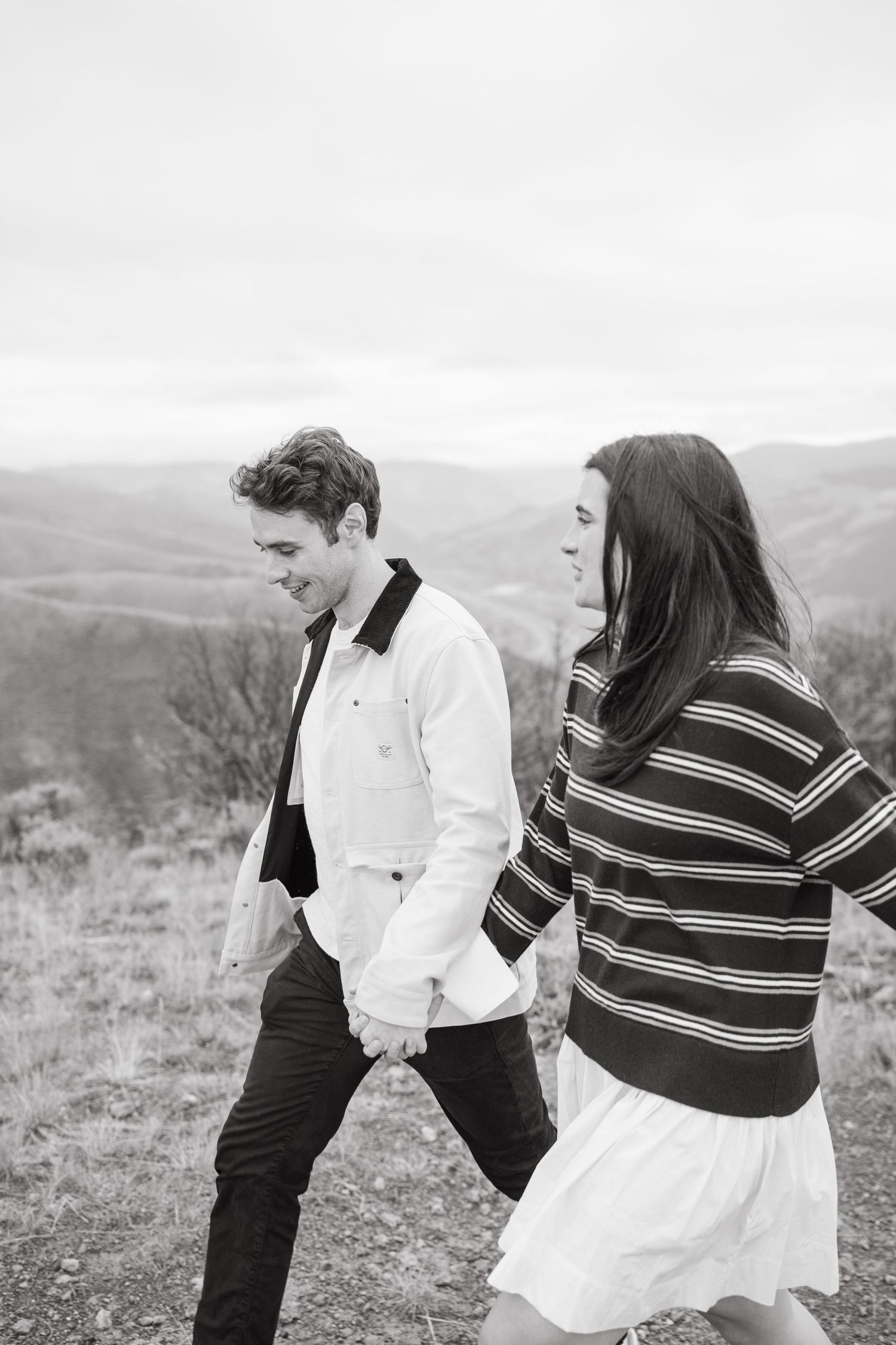 A couple walks hand in hand along a hilltop trail, with rolling mountains in the background on an overcast day.