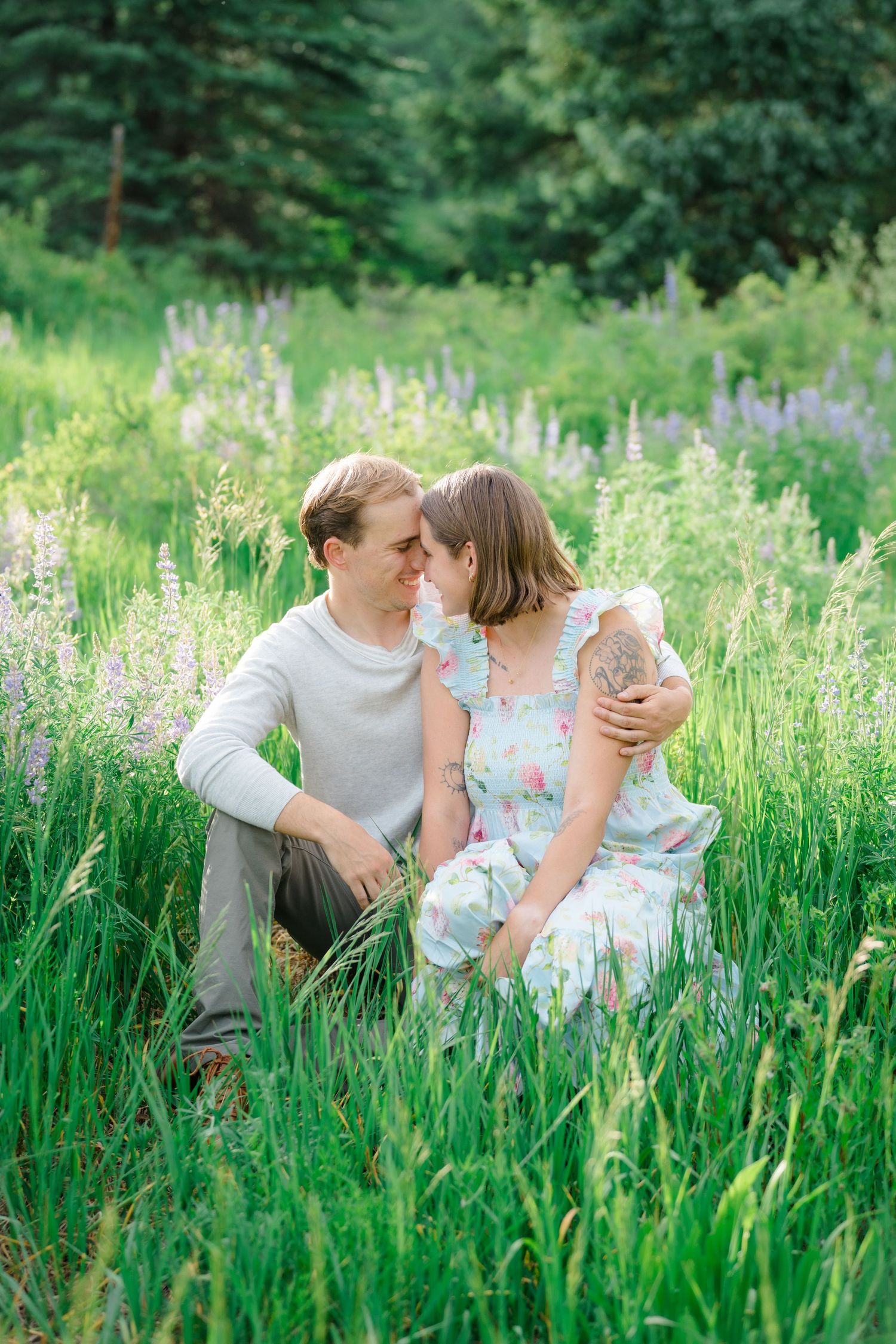 A couple shares tender moments while sitting in a lush green meadow filled with tall grass and wildflowers.