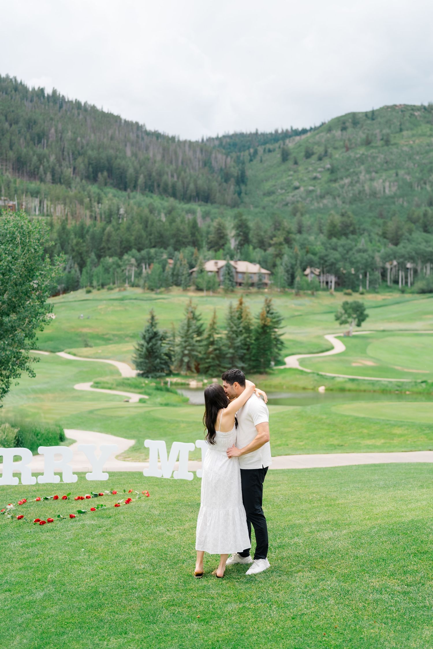 A romantic proposal scene unfolds on a scenic golf course with rolling green hills and mountains in the background.