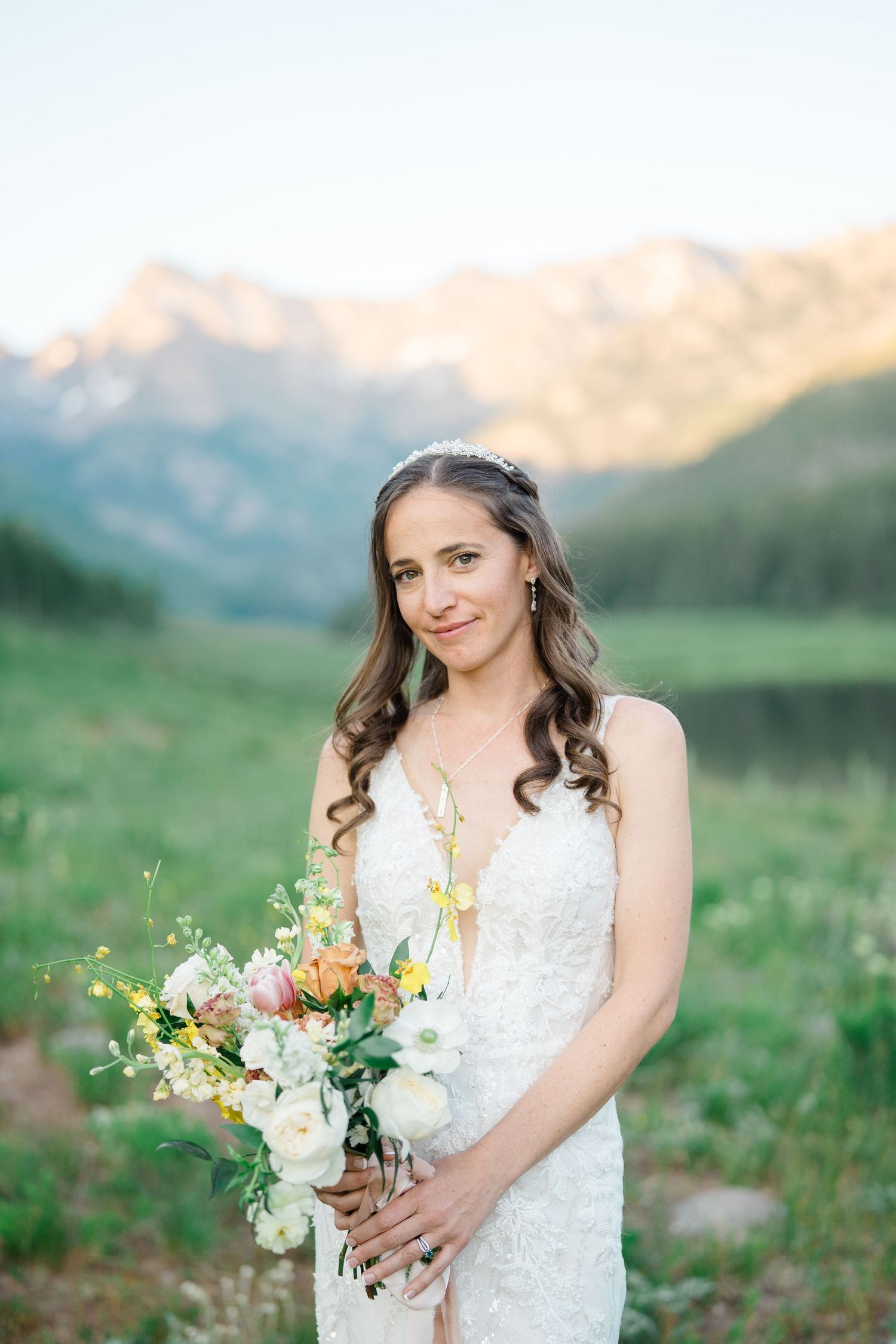 Bride in lace dress holds colorful wildflower bouquet against mountain landscape backdrop.