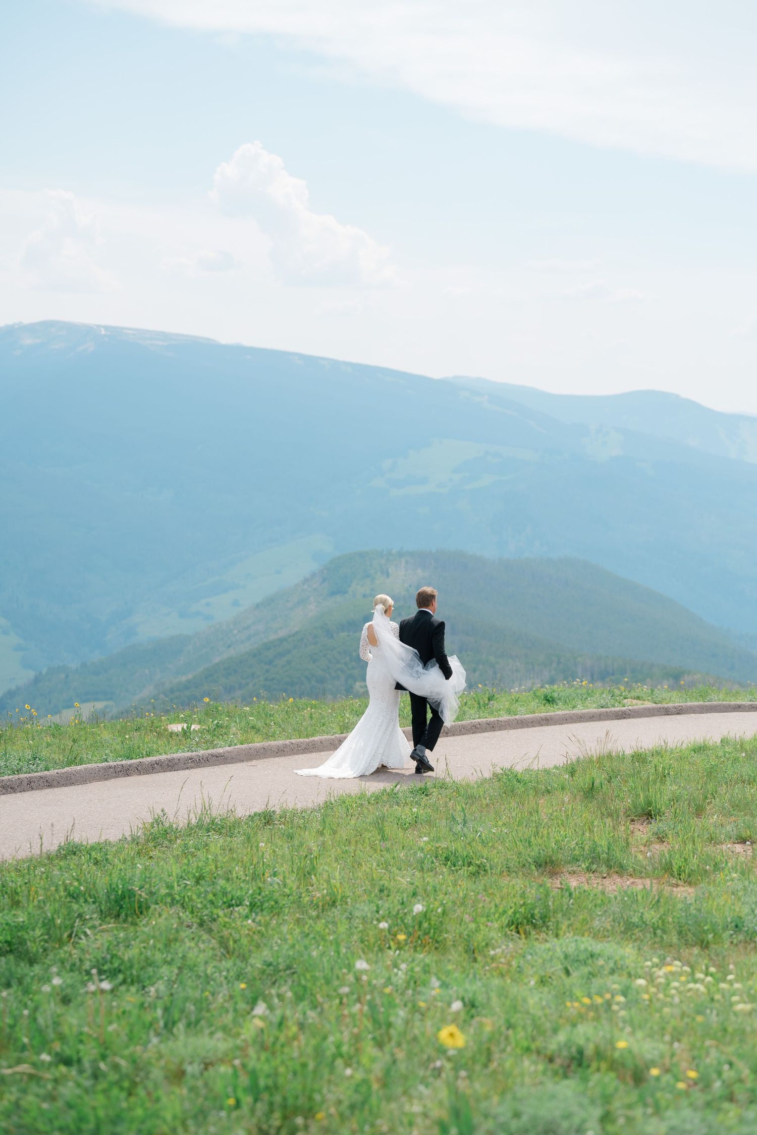 A couple in wedding attire walks along a scenic mountain path surrounded by wildflowers and rolling hills.