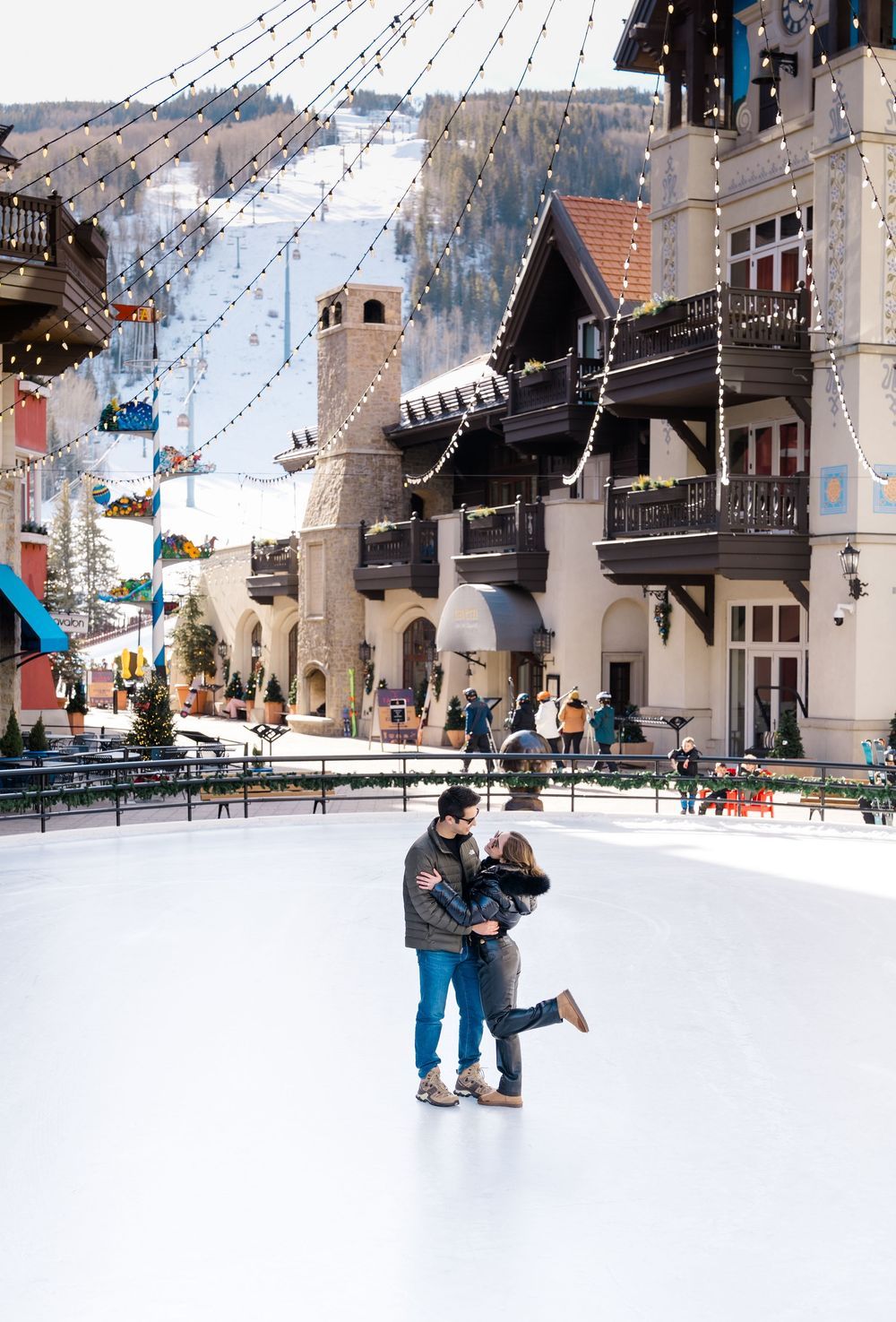Ice skaters glide across an outdoor rink beneath twinkling lights with European-style buildings in the background during vail proposal.