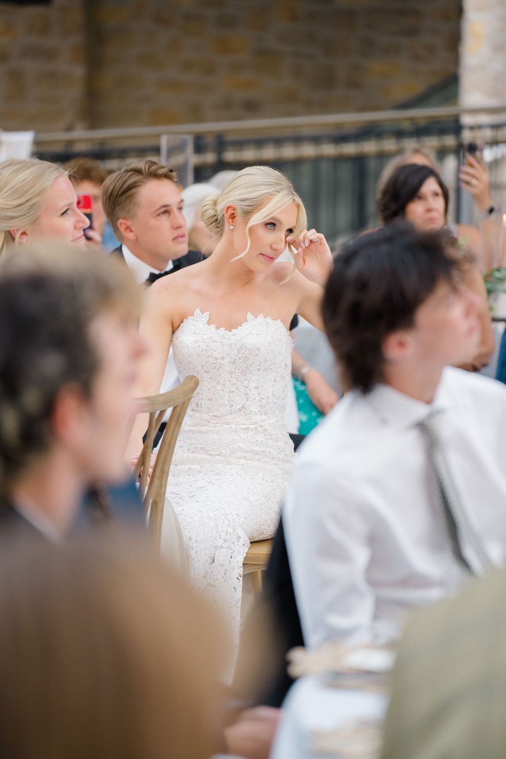 Blonde bride in a white lace strapless gown sits among wedding guests at an outdoor reception, smiling and touching her hair.