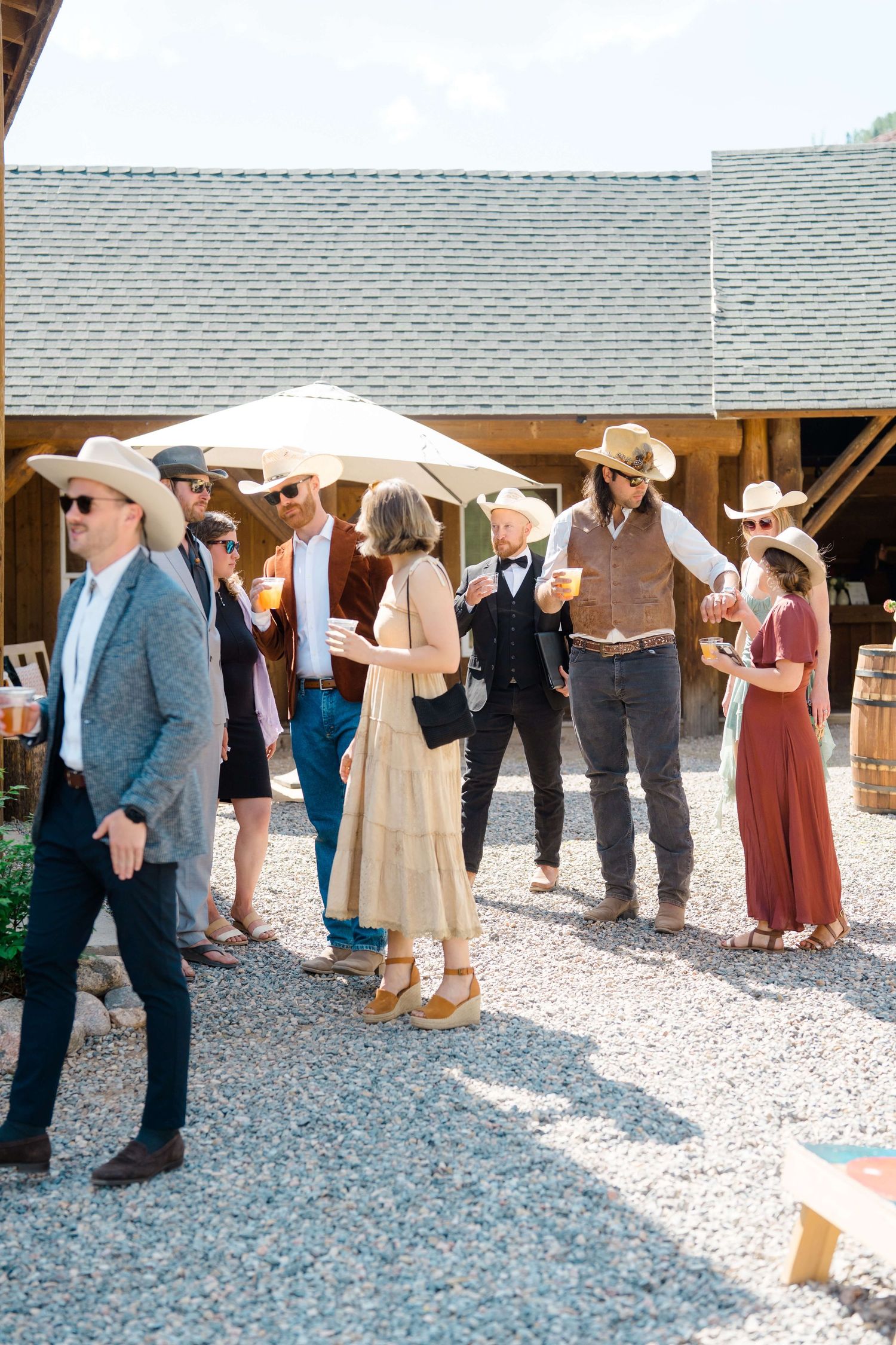 Guests in western attire with cowboy hats mingle outdoors at a rustic barn wedding reception on a sunny day at Piney River Ranch.