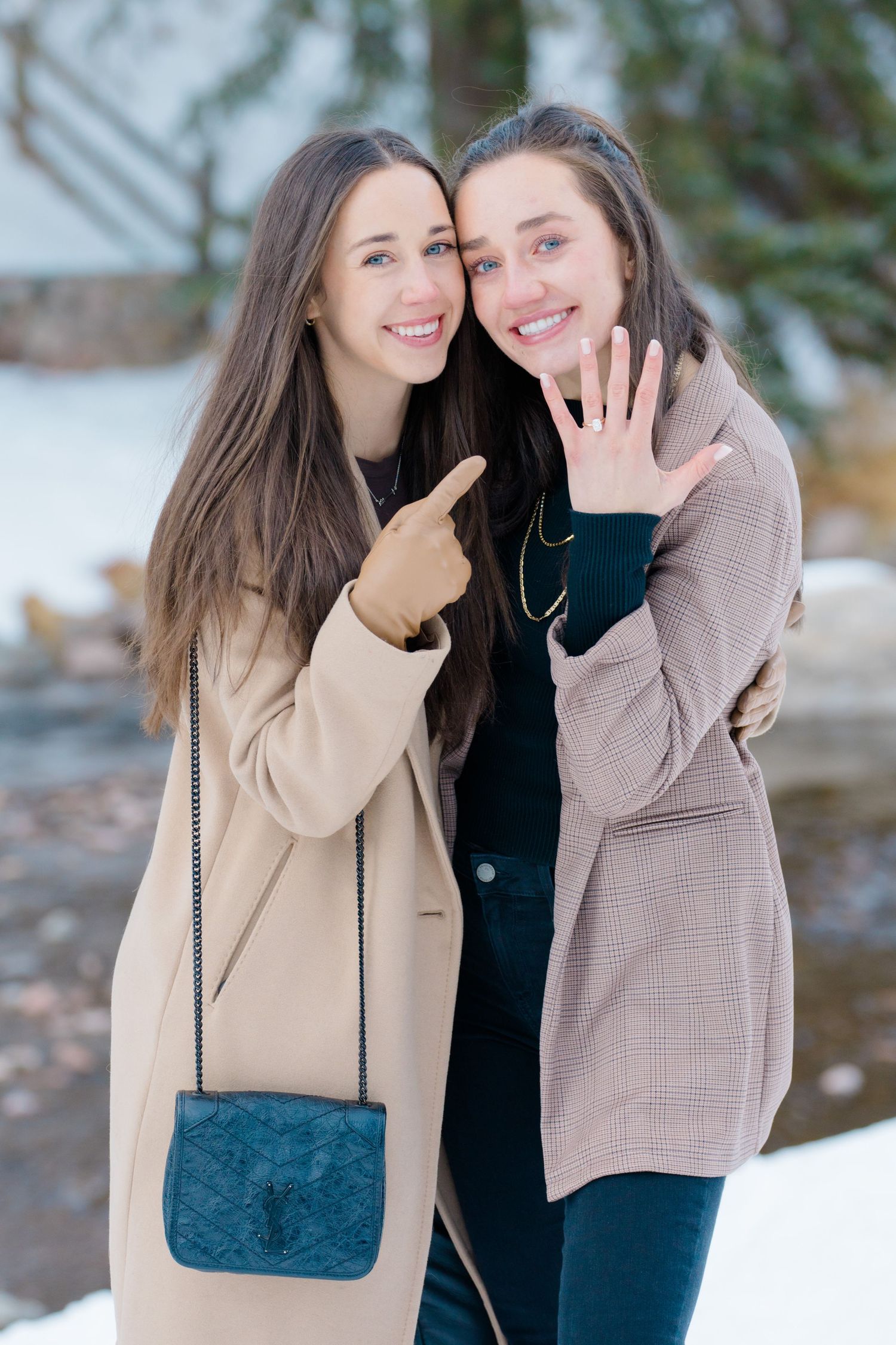 Two friends in winter coats celebrate an engagement outdoors in the snow, with one pointing to a ring.