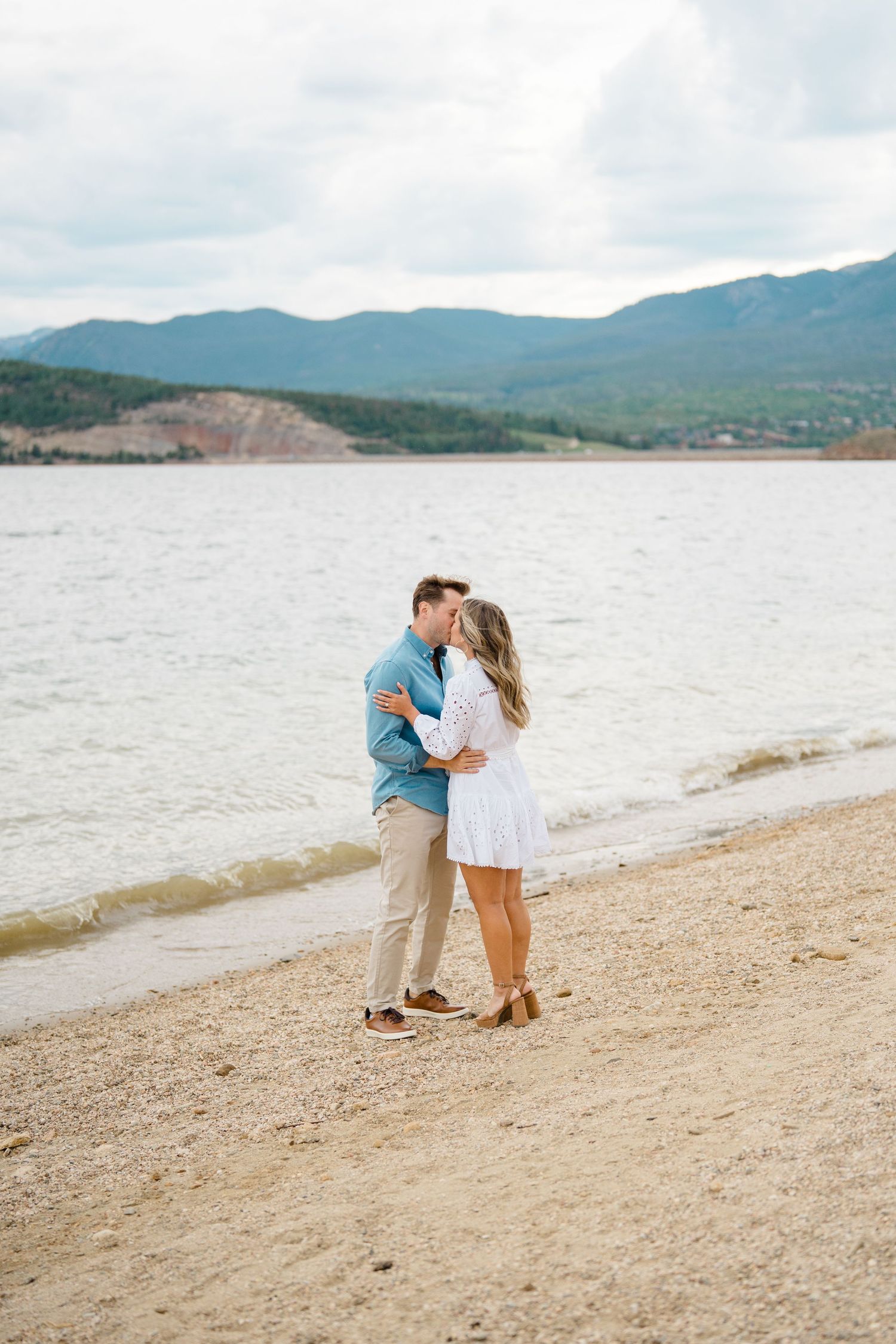 A couple shares a romantic moment on a sandy beach with mountains and lake in the background.