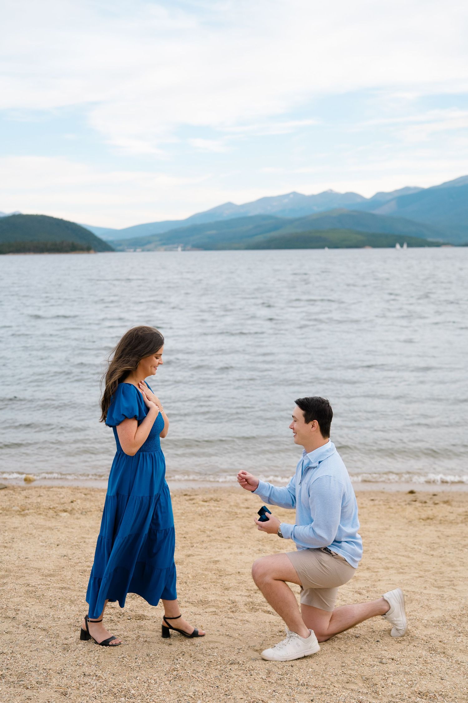 A couple shares a romantic proposal moment on a scenic beach with mountains and lake in the background.