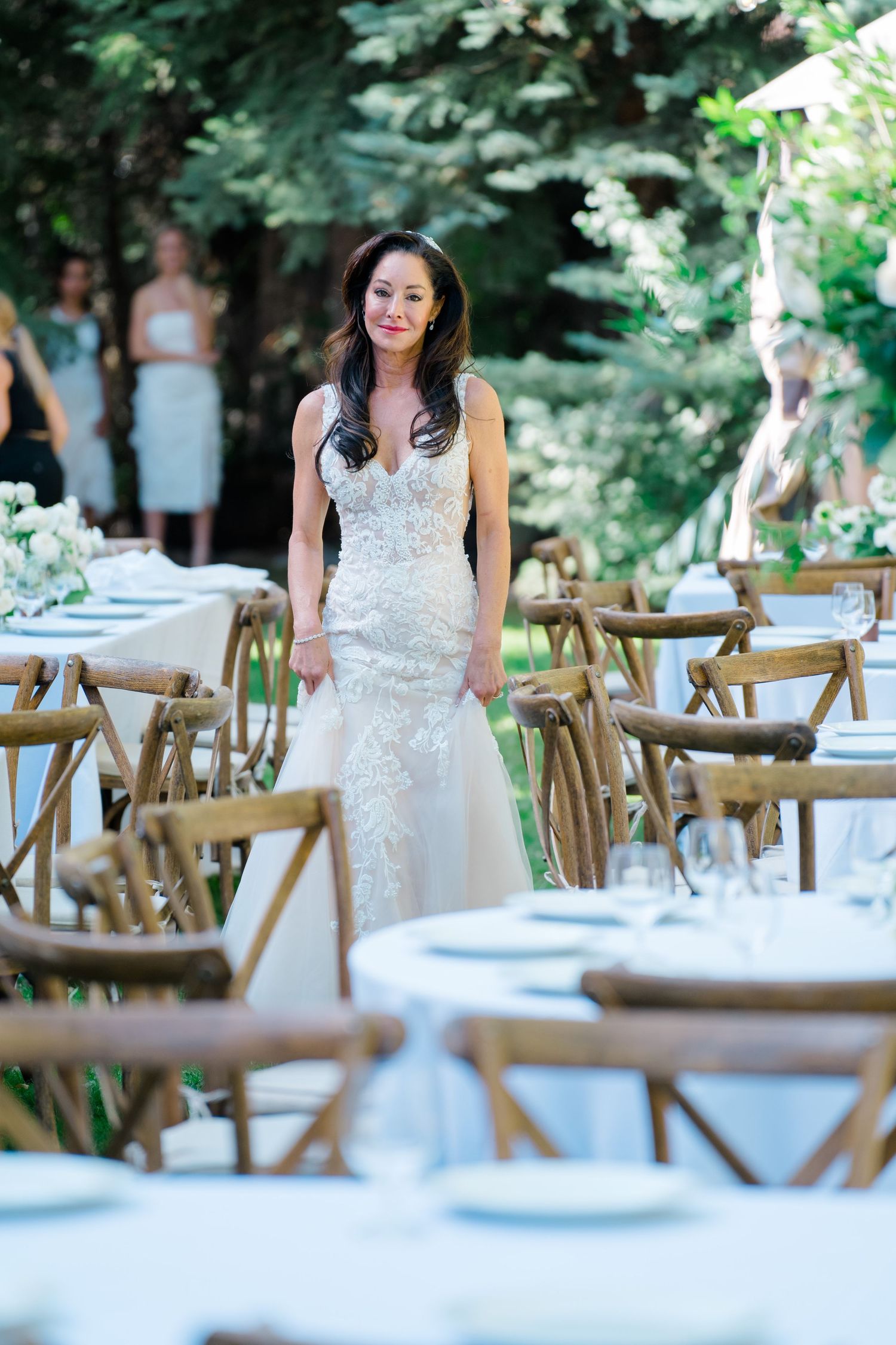 A wedding reception setup outdoors with wooden chairs and white tablecloths in a garden setting.