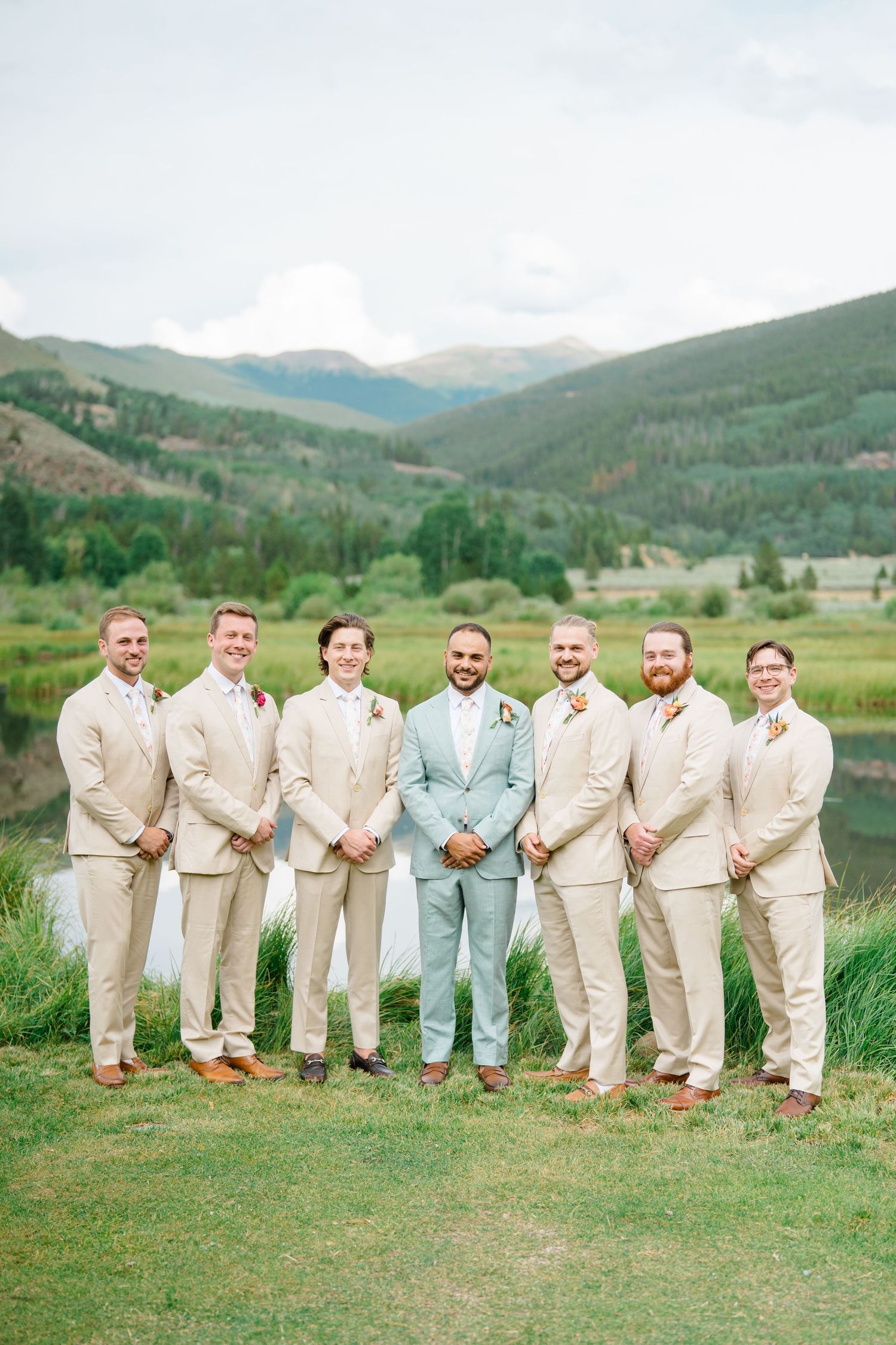 Wedding party in beige suits and one blue suit posing on grassy hill with mountain backdrop.