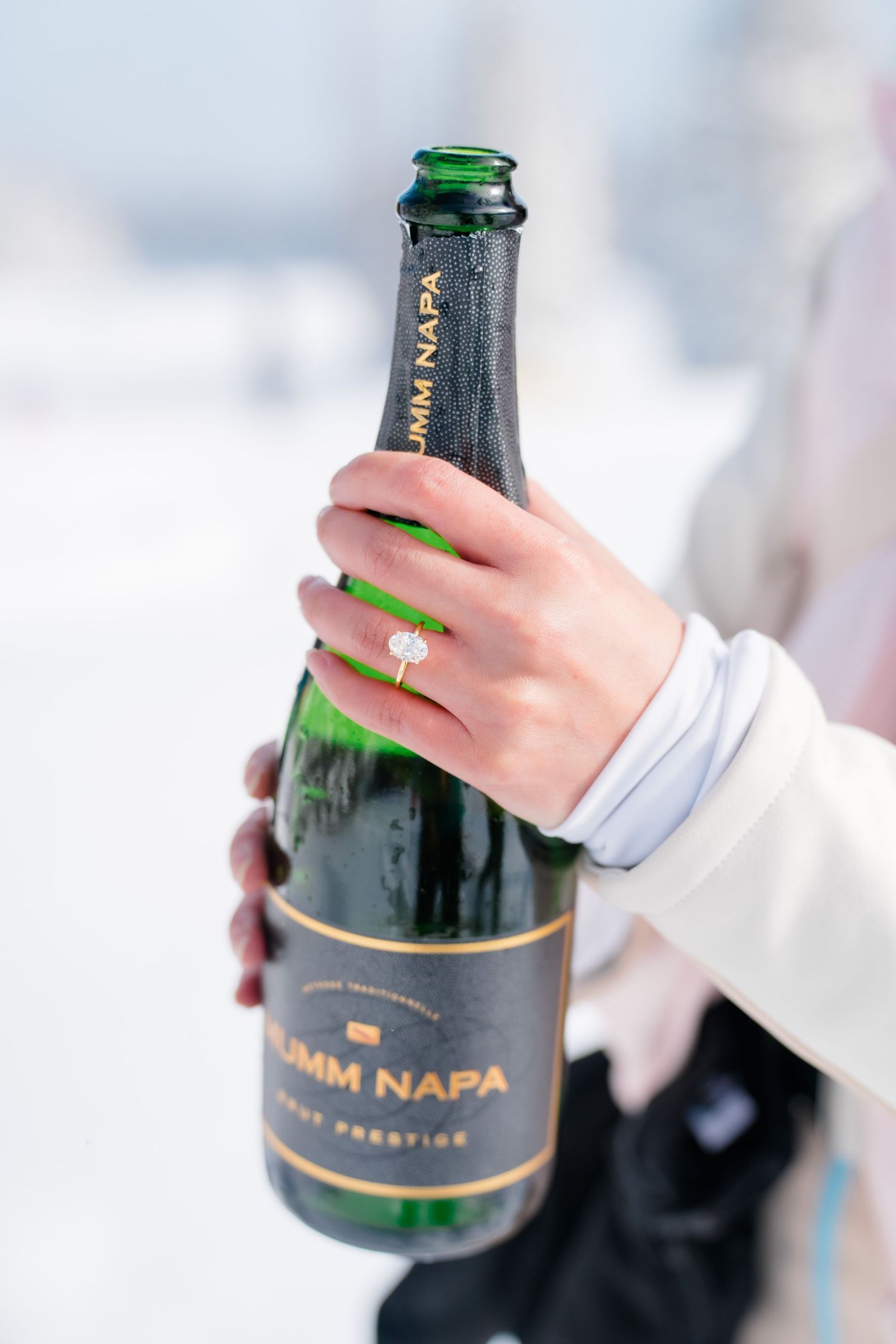 A hand wearing a white sleeve and emerald ring holds a bottle of Domain Napa wine against a blurred background.