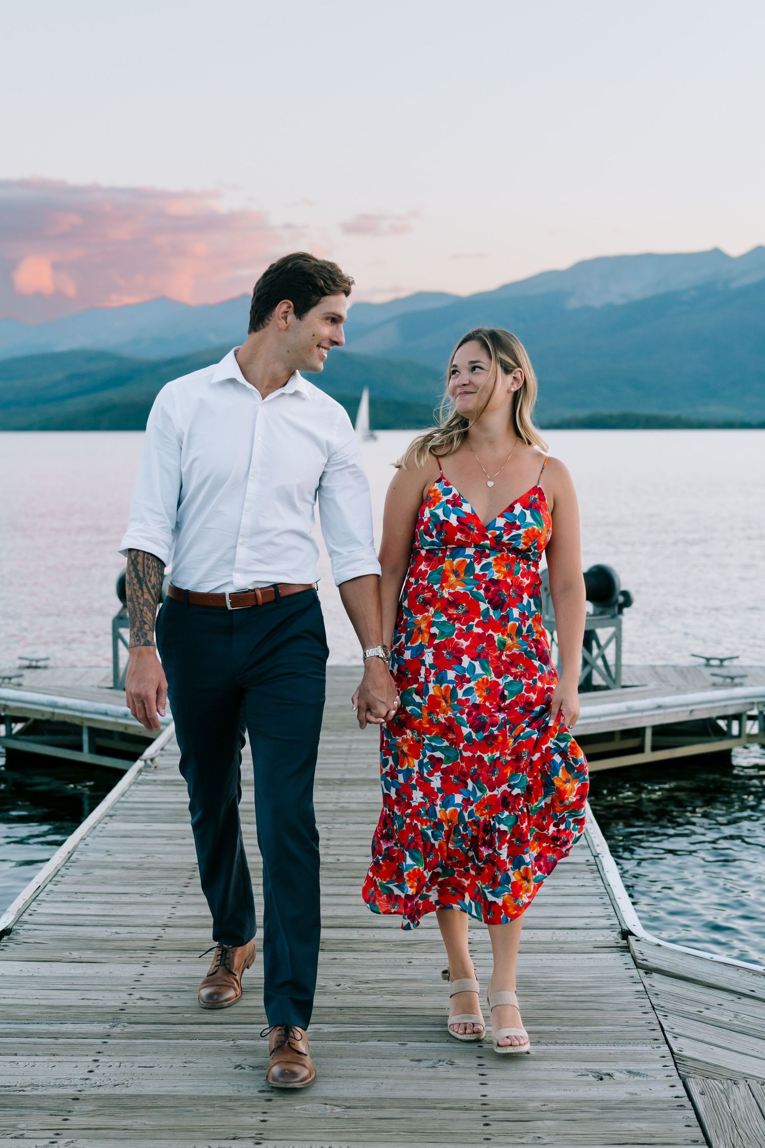 A couple sits together on a lakeside dock at sunset with mountains in the background.
