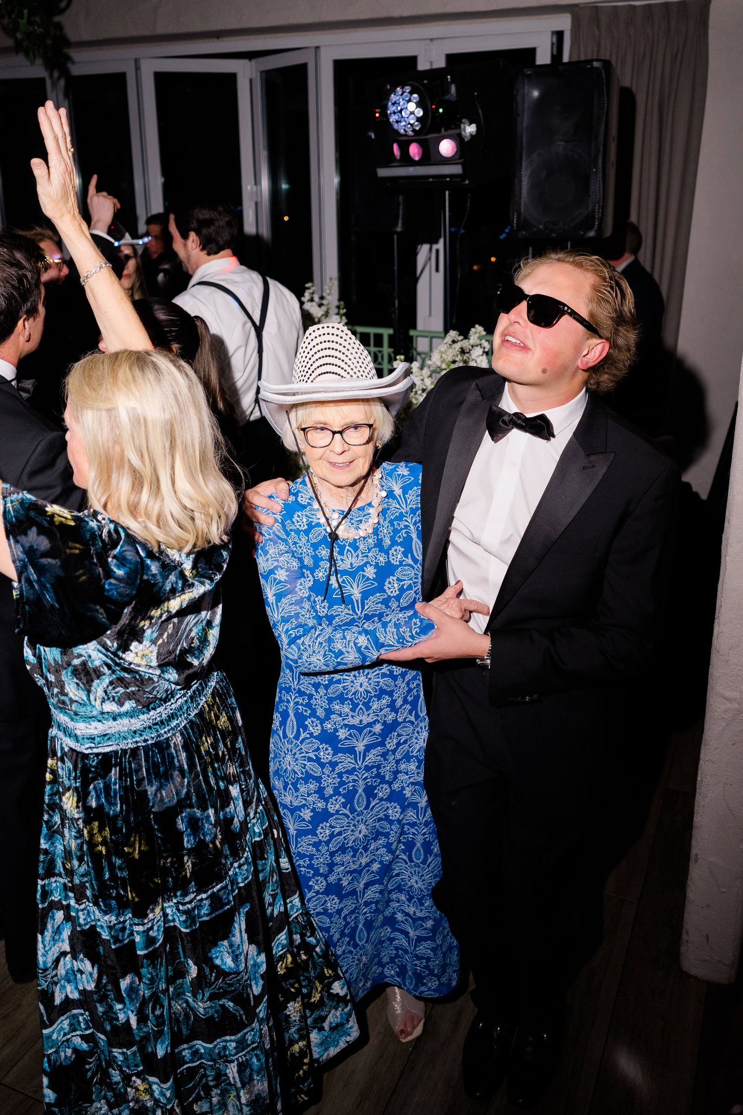 Elderly woman in blue floral dress and white cowboy hat dancing with a man in tuxedo and sunglasses at a wedding reception.