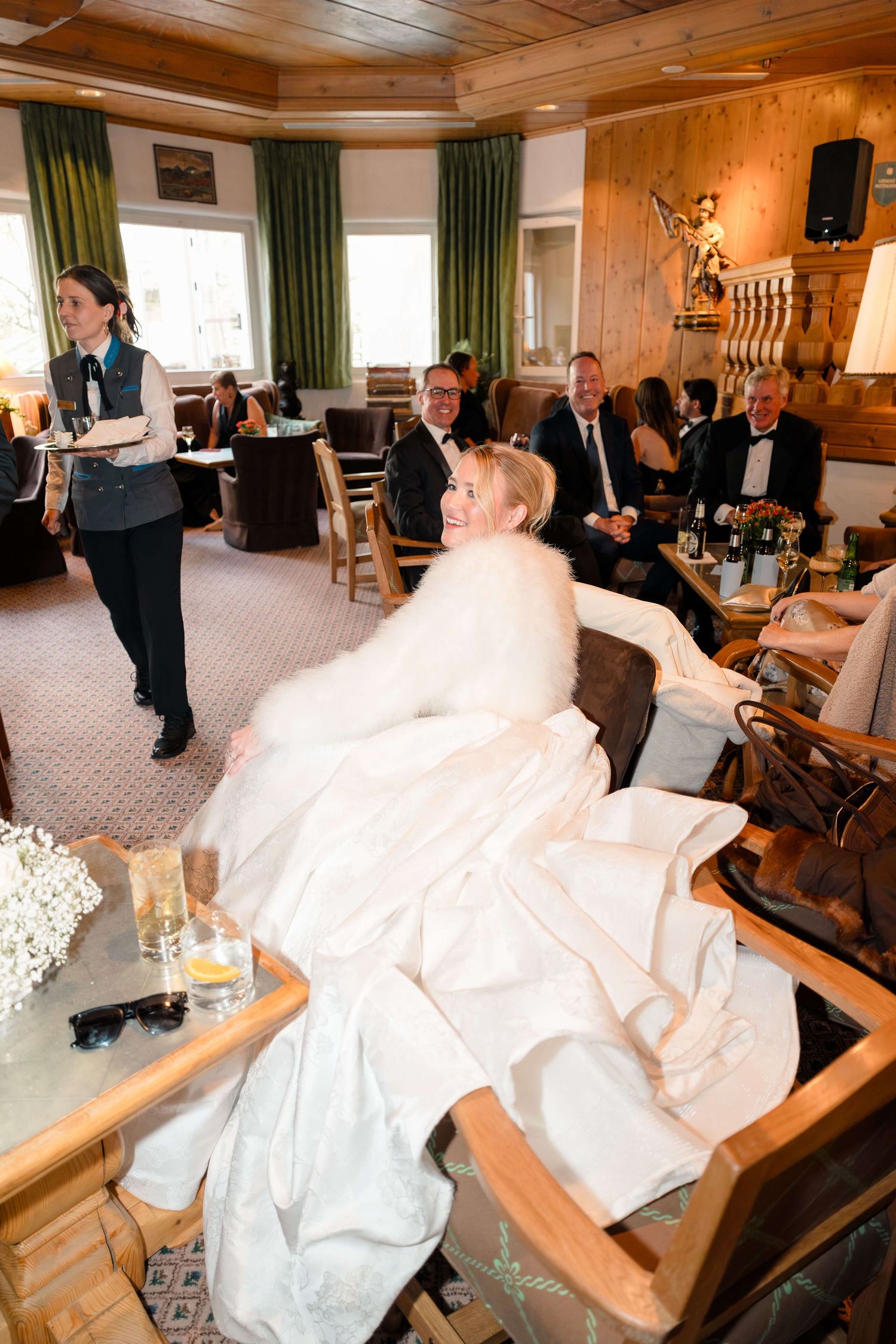 Bride in white gown with fur wrap seated at wedding reception in a cozy wood-paneled venue.
