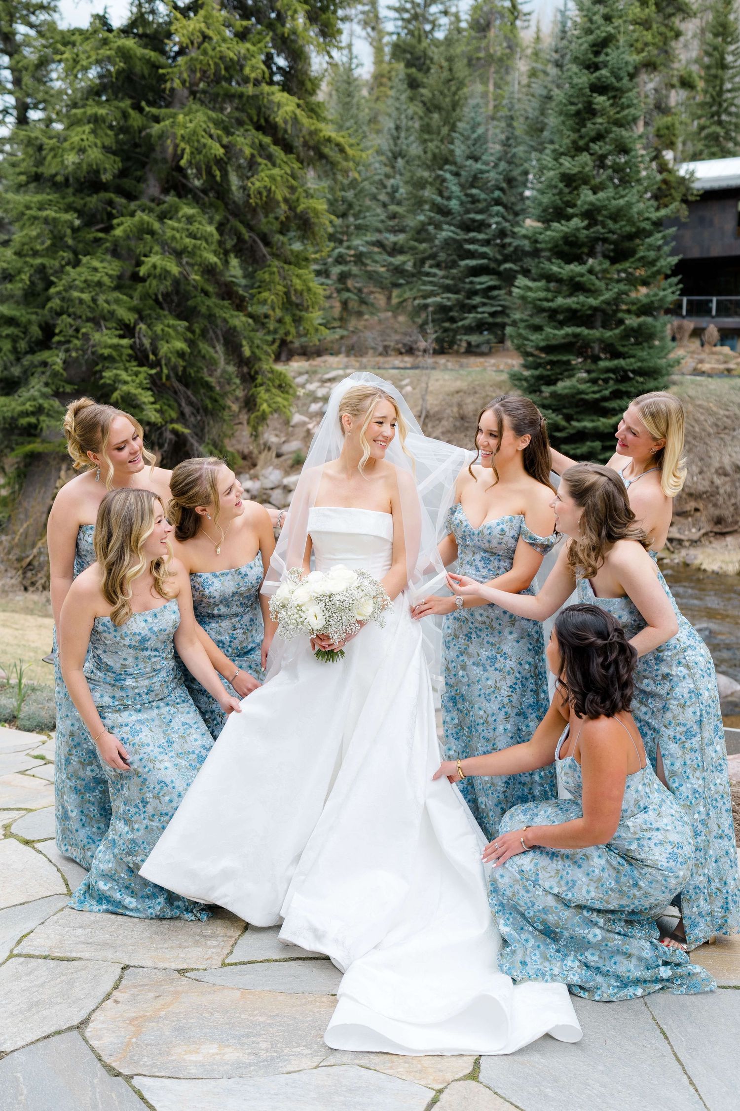 Bride in white gown with bridesmaids in blue floral dresses on stone patio surrounded by pine trees at Vail Sonnenalp Wedding.