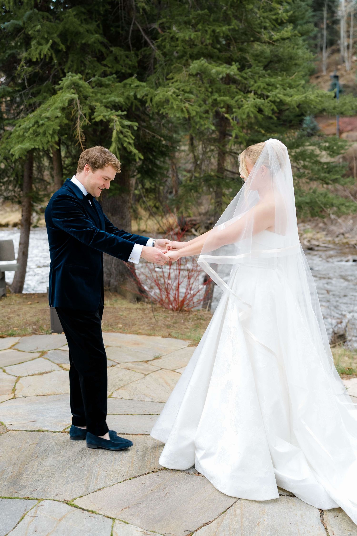 Groom in navy suit holds bride's hands outdoors on stone patio surrounded by evergreen trees.