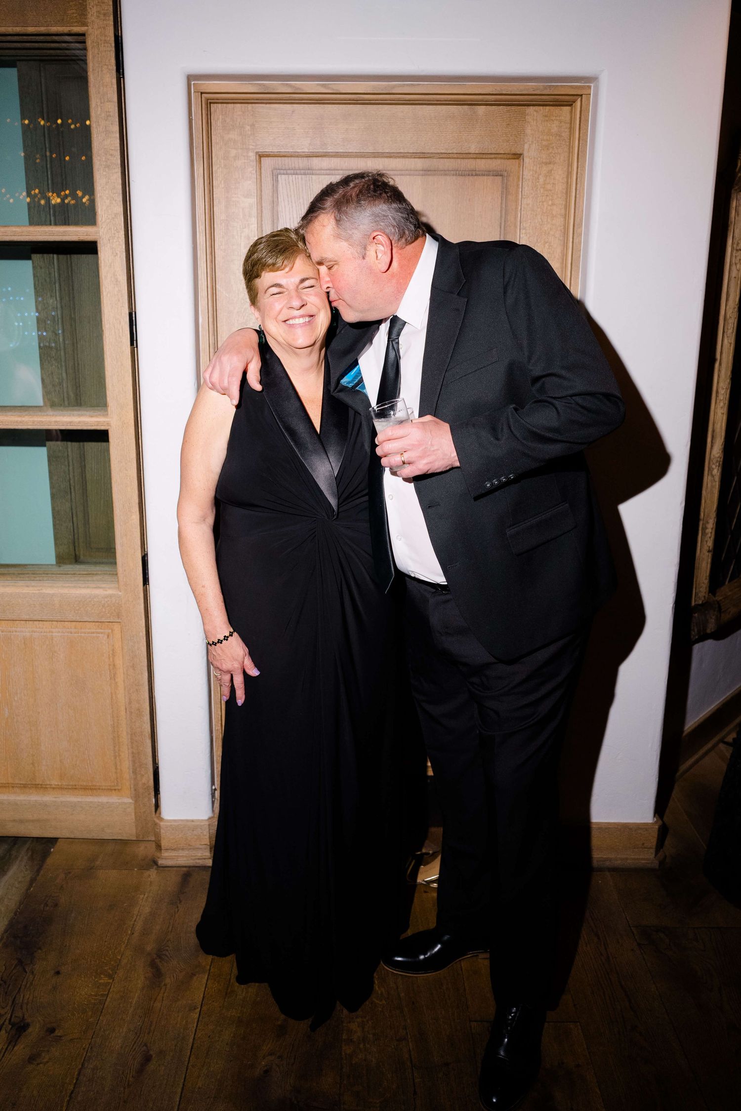 Couple in formal black attire sharing a tender moment at an elegant evening event, man kissing woman's cheek.