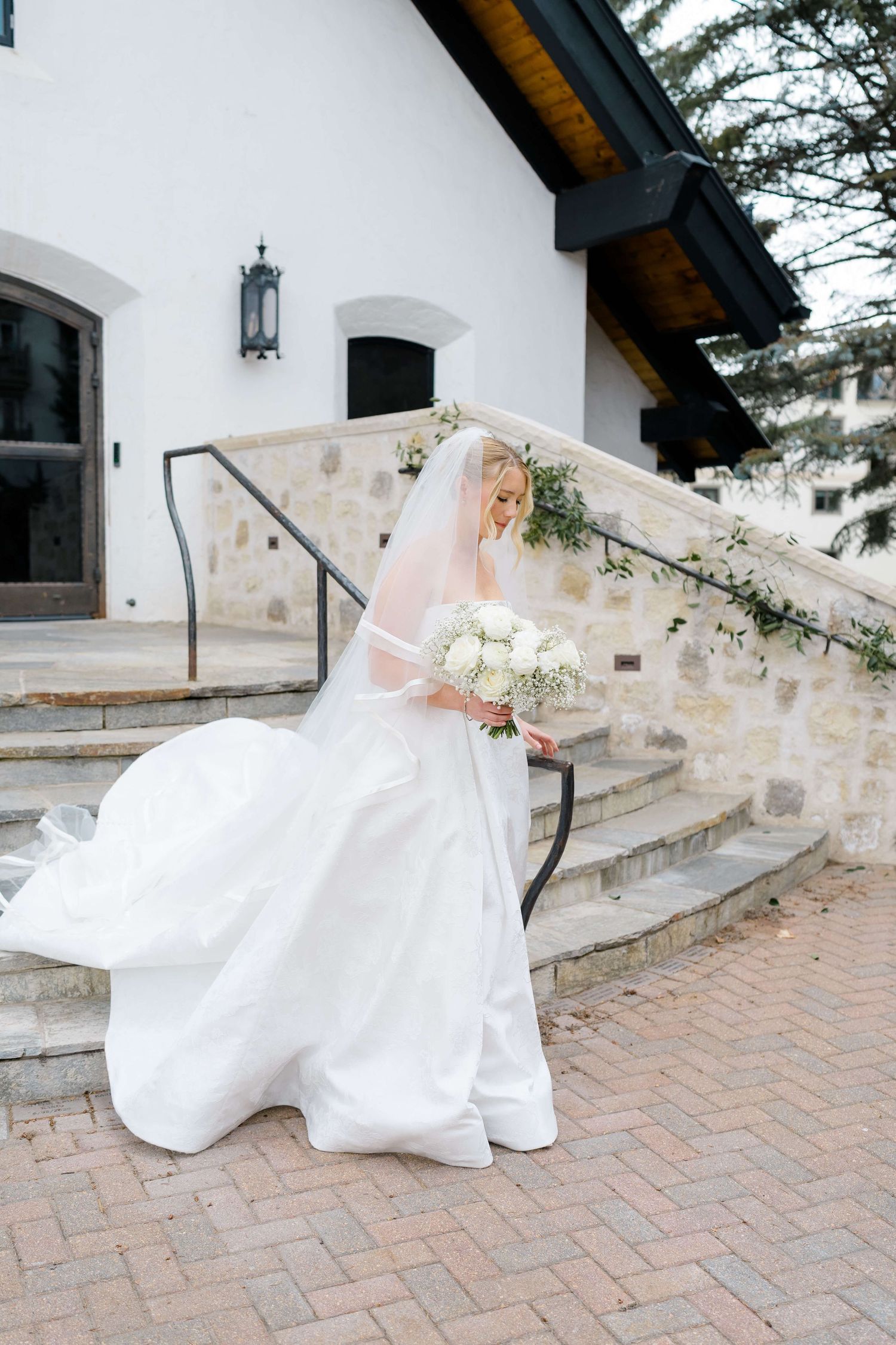 Blonde bride in flowing white ball gown and veil holds white bouquet on stone chapel steps.