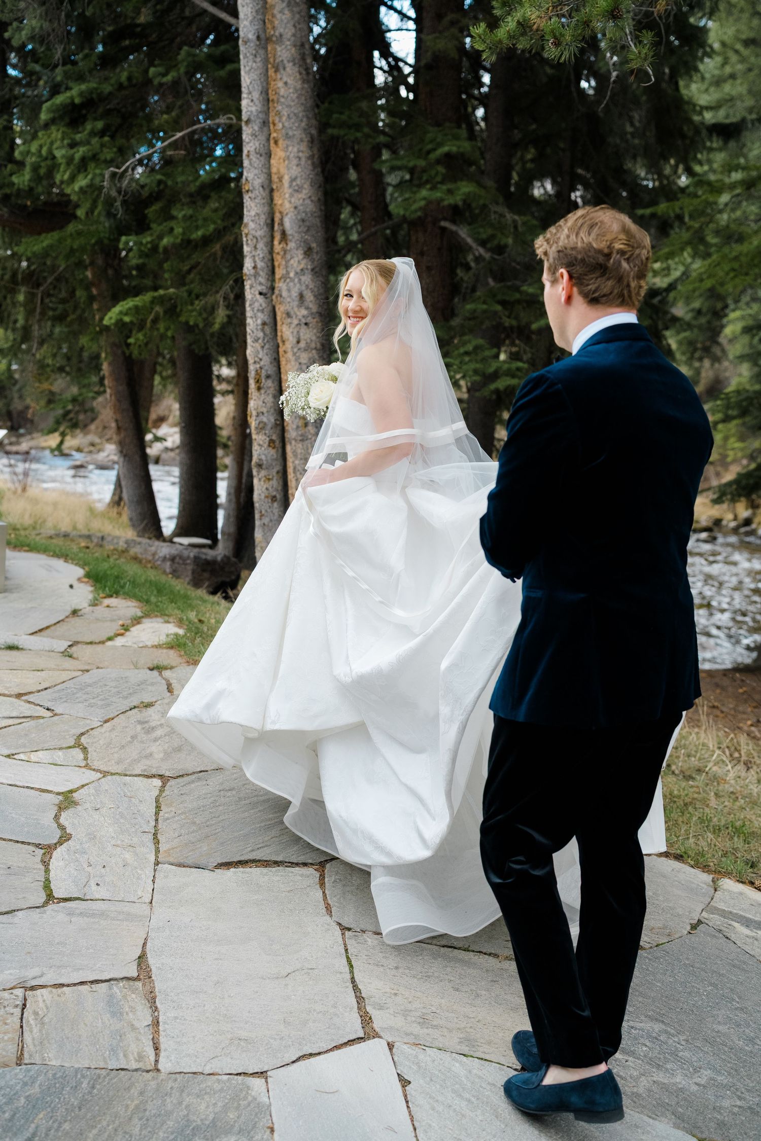 Bride in white gown and veil walks with groom in navy suit along stone path surrounded by pine trees taken by vail wedding photographer