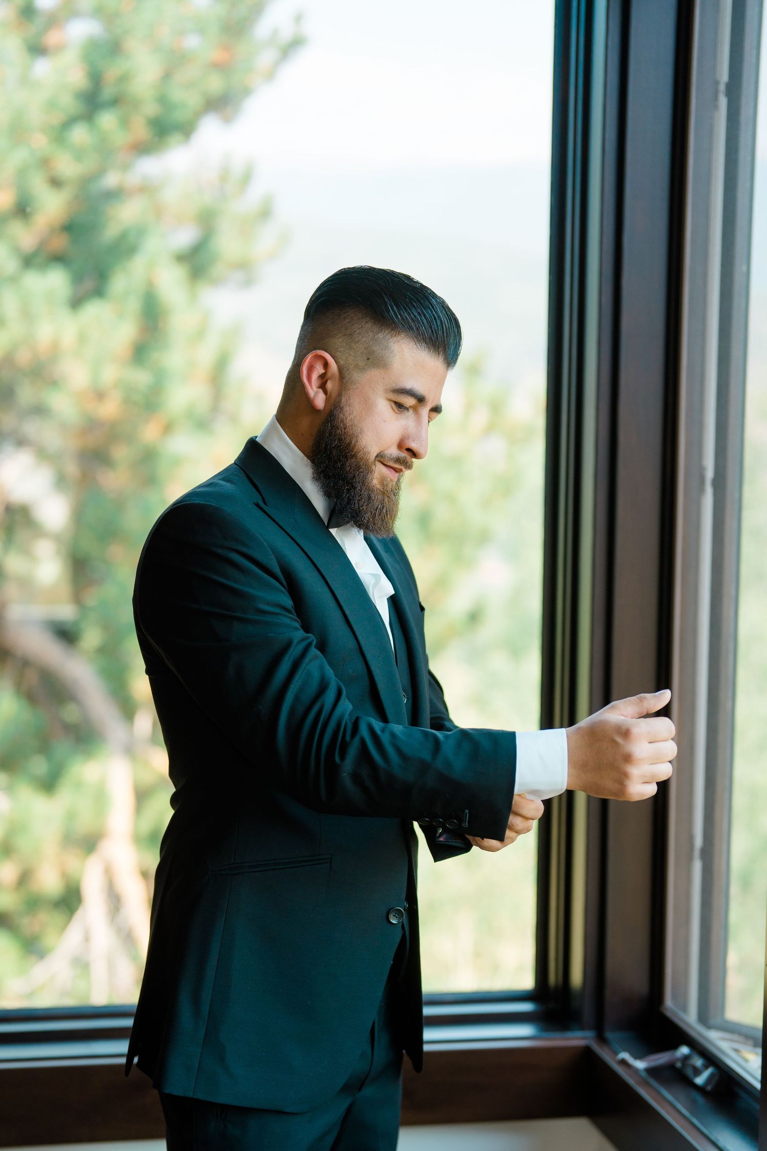 A groom in a dark suit adjusts his cufflinks while standing by large windows in a sequence of candid shots during Breckenridge wedding.