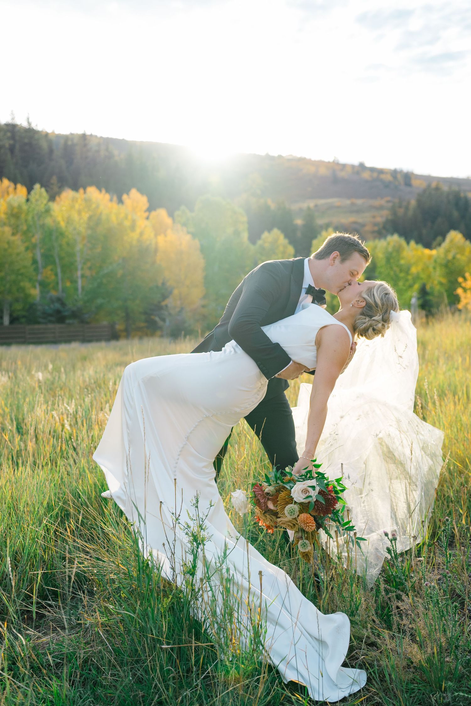 Groom dips bride for a kiss in a sunlit meadow with autumn trees, bride holding a colorful bouquet in a white gown.