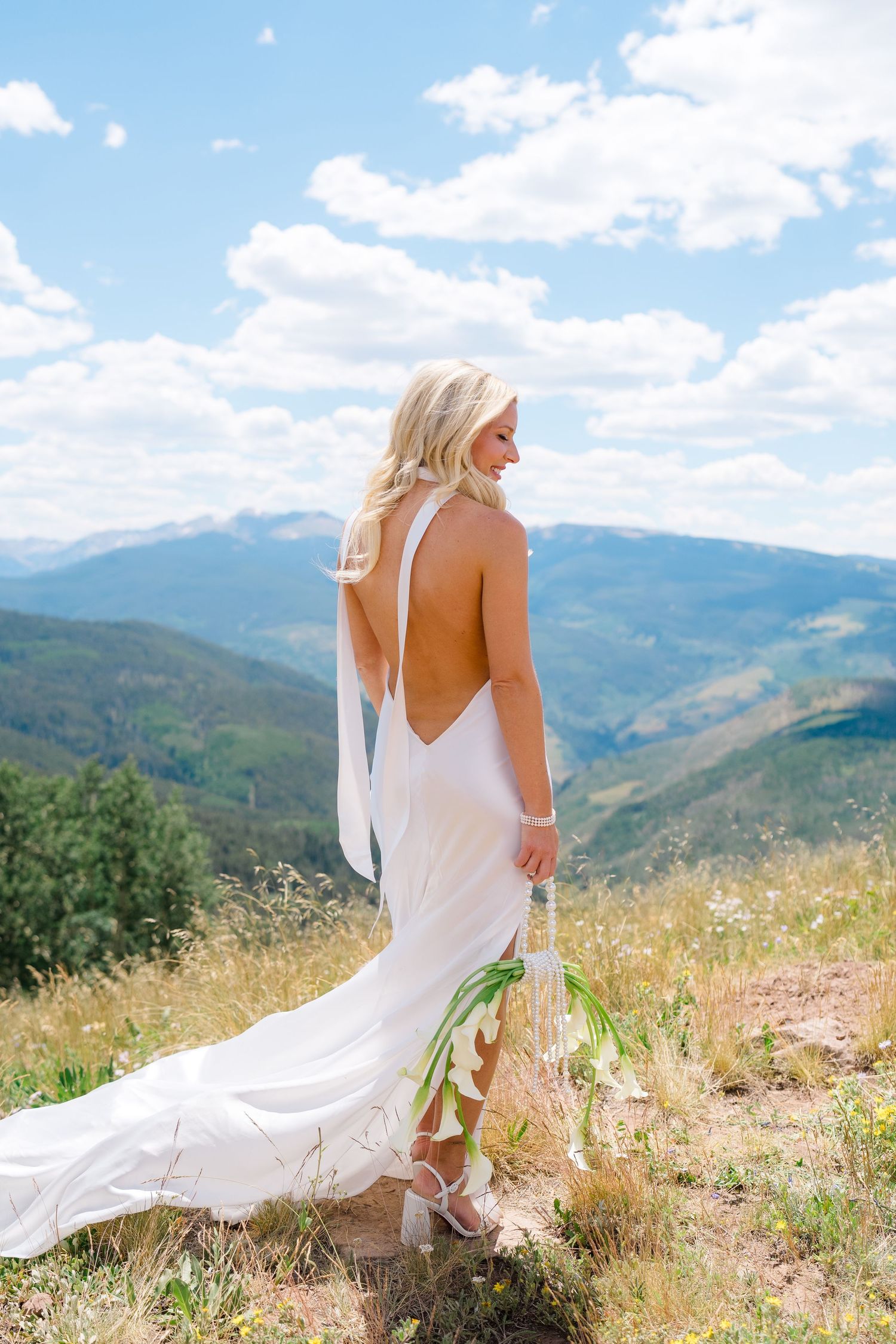 Blonde bride in backless white halter gown holds bouquet on mountain overlook under blue sky.