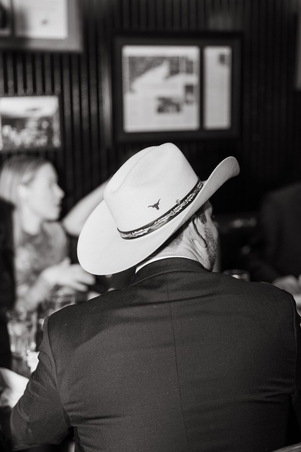 Black and white photo of a man wearing a cowboy hat with longhorn emblem at a bar event.