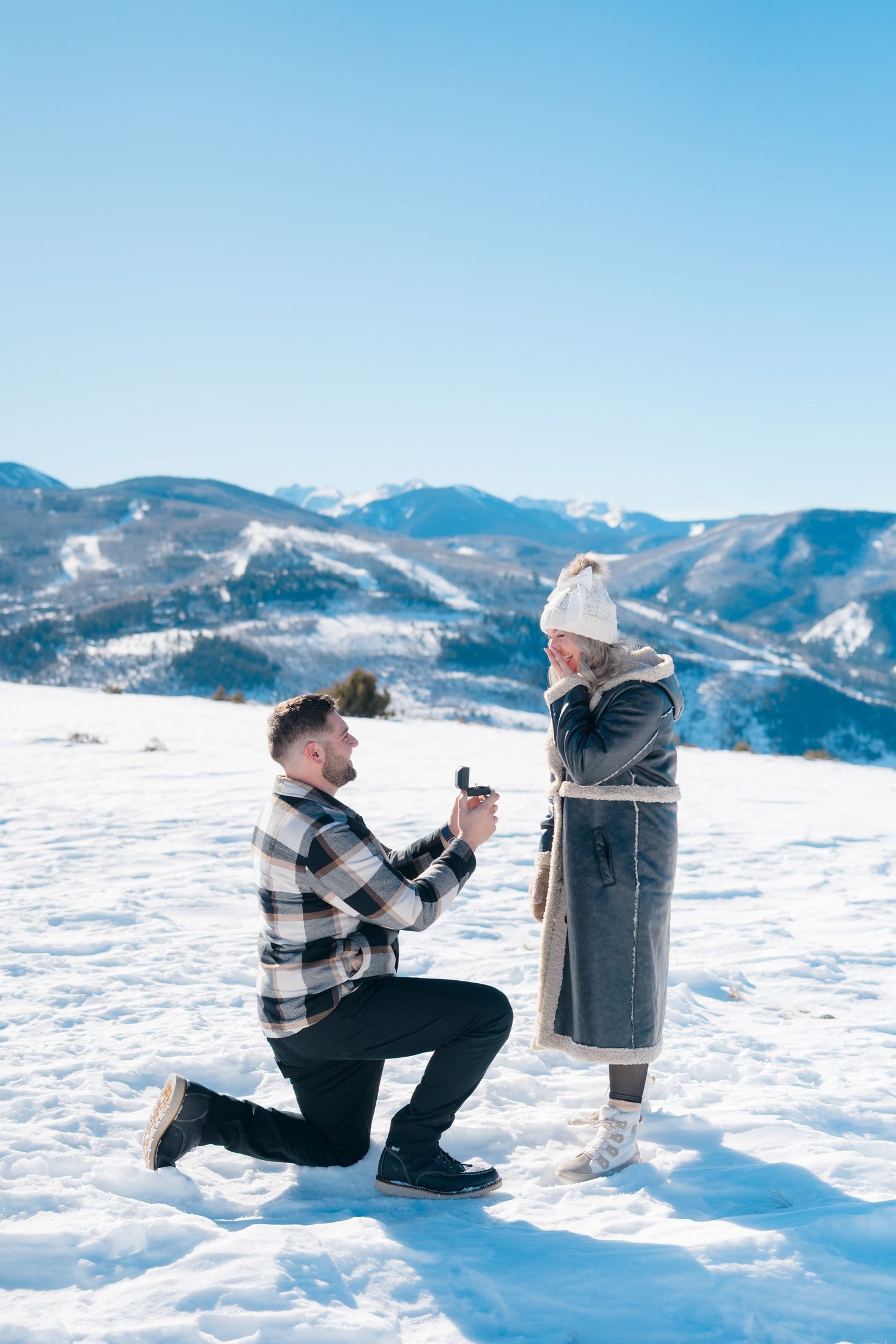 A snowy mountain proposal scene unfolds with figures kneeling in fresh powder against a bright blue sky during vail proposal.