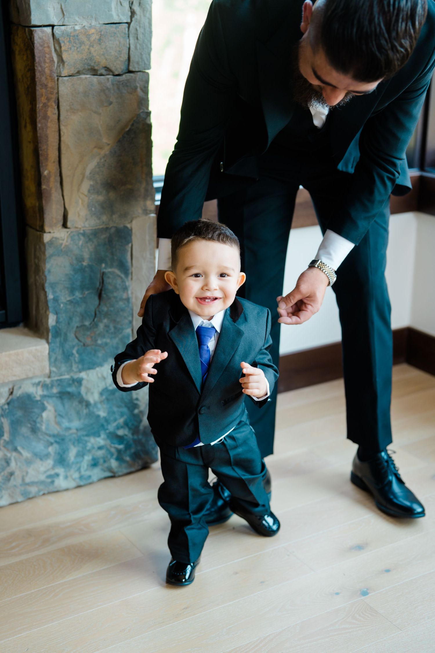 A small child in a suit and tie stands smiling while an adult in matching formal attire helps adjust outfit during Breckenridge wedding.