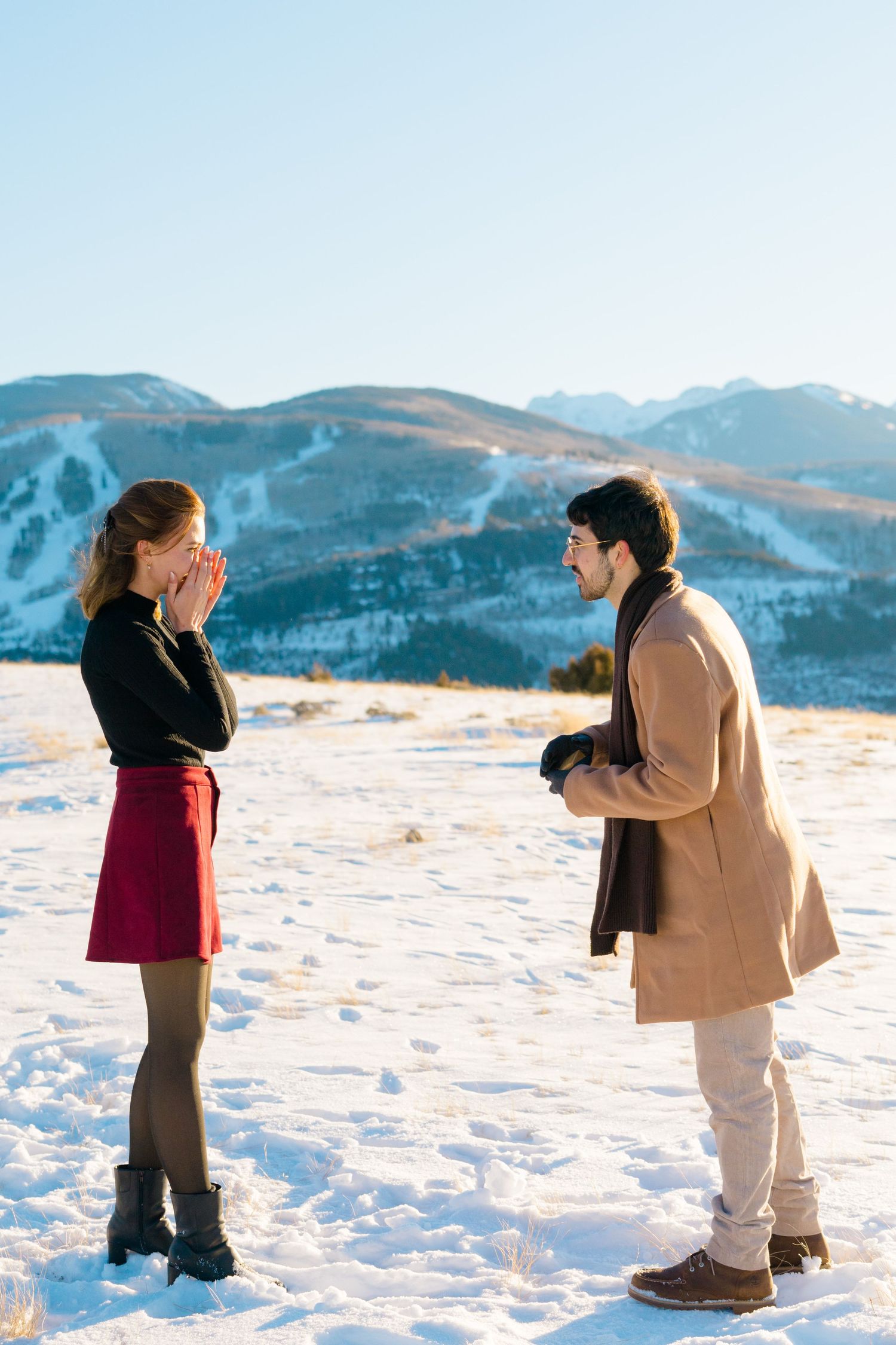 A couple shares a romantic winter proposal moment on a snowy mountain overlook with scenic peaks in the background.