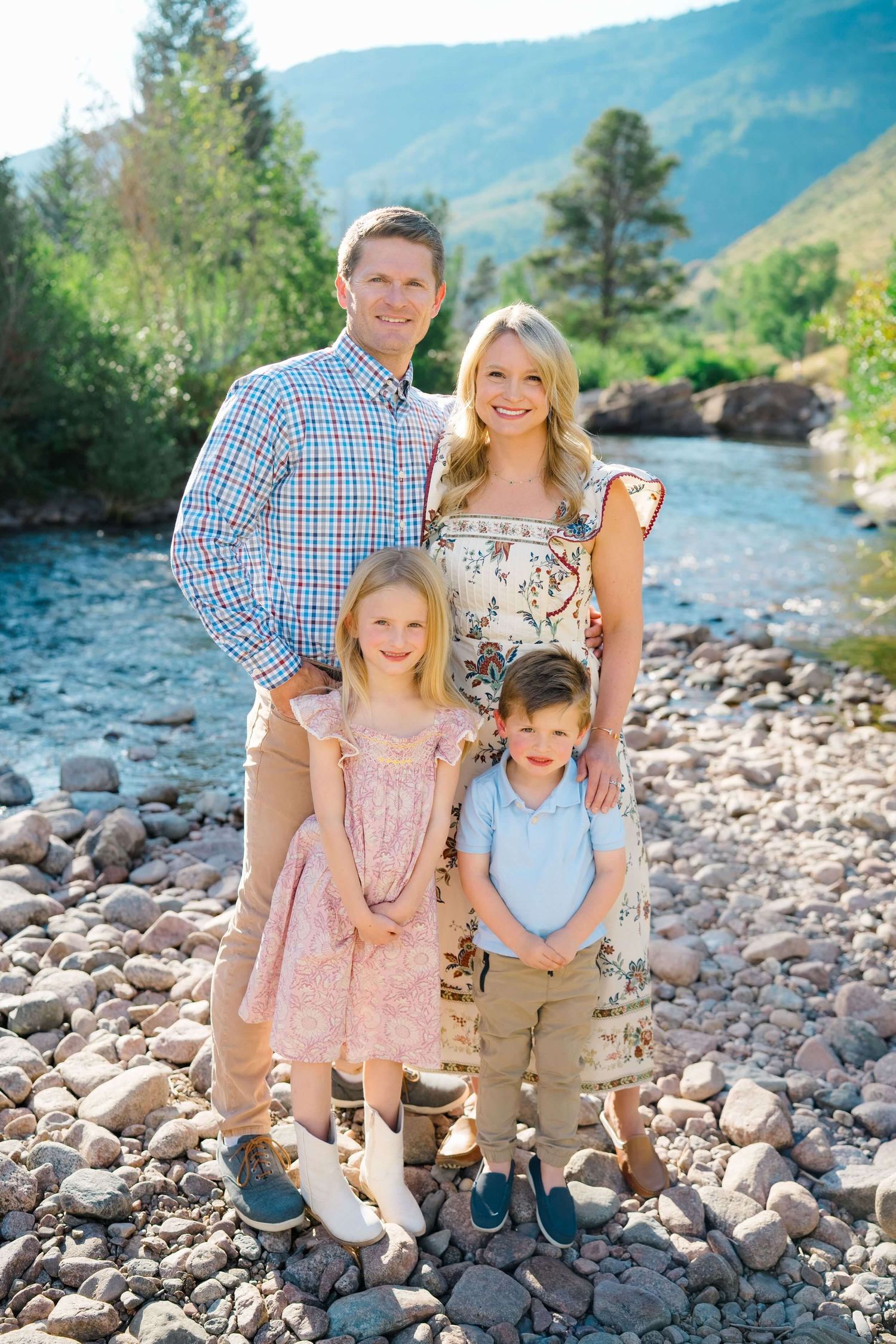 Family of four poses together on rocky riverbank with mountain landscape and flowing stream in background.
