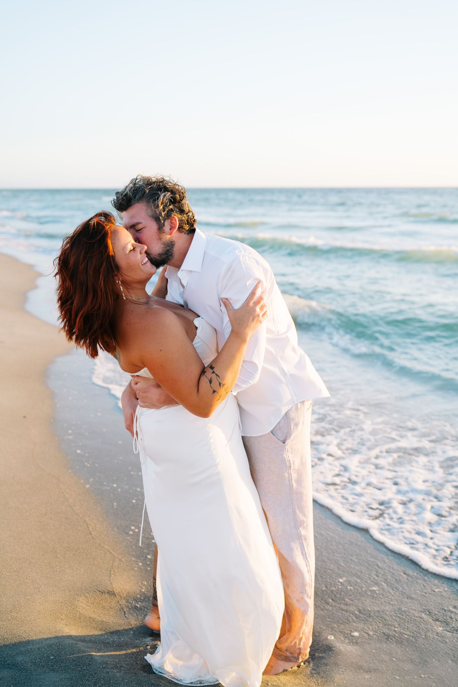 A romantic couple in white embraces on a sunny beach with ocean waves in the background.