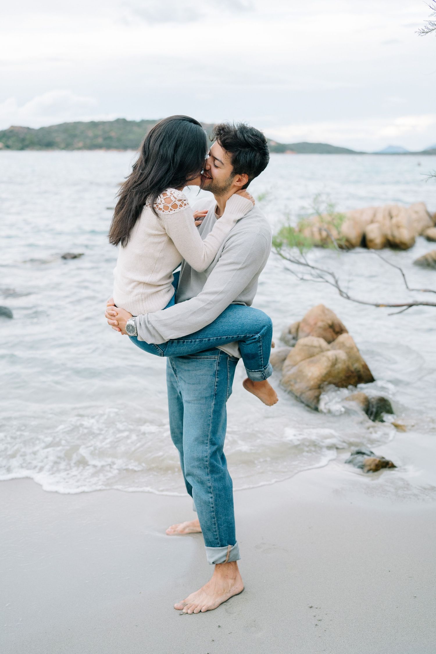 A couple embraces and kisses on a rocky beach with waves and mountains in the background.
