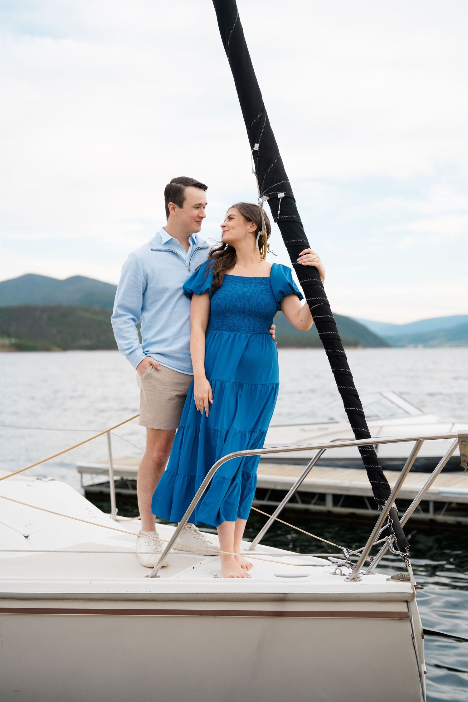 A couple stands together on a sailboat deck with mountains and lake in the background.