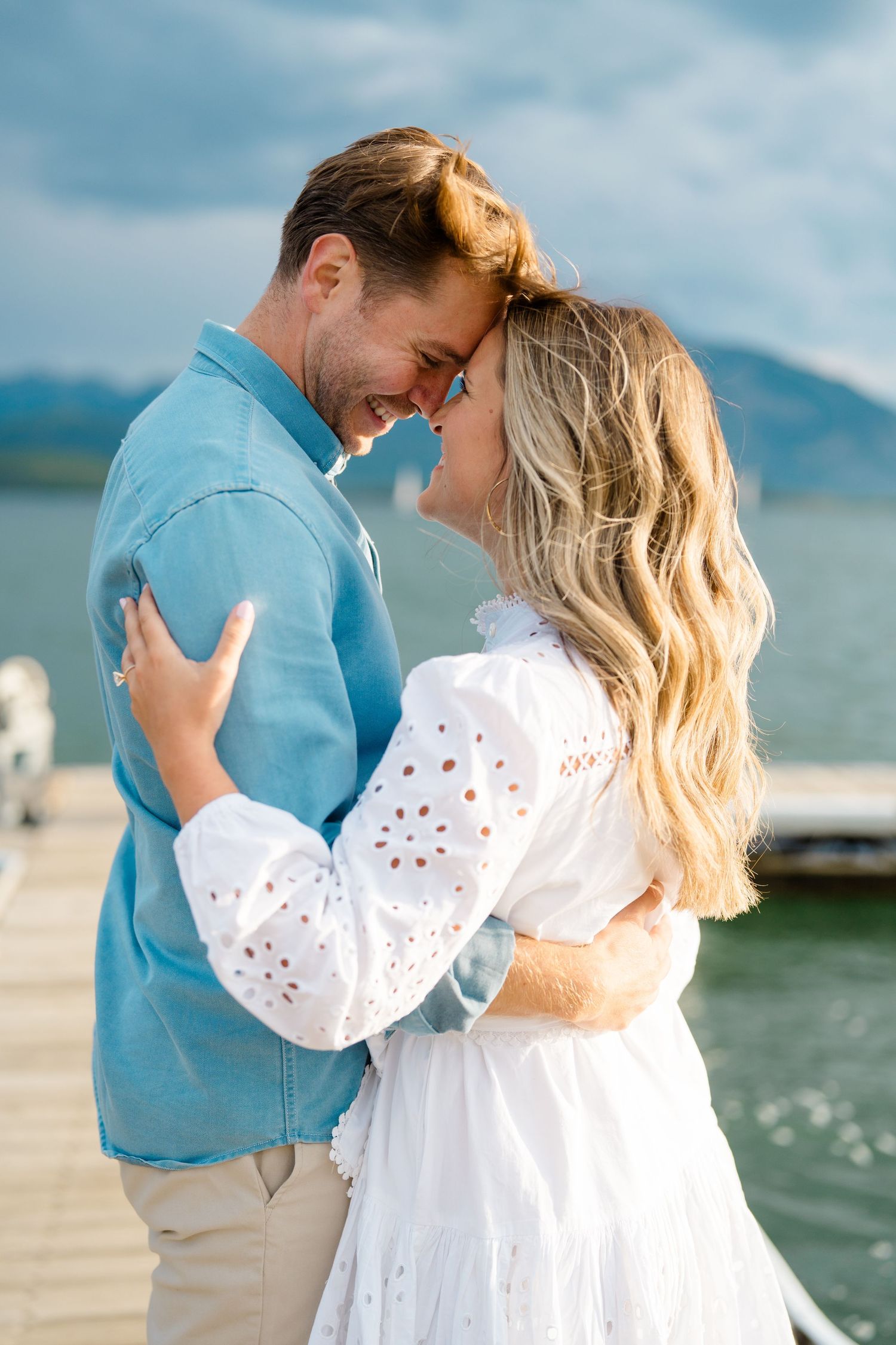 A couple shares an intimate moment on a lakeside dock with mountains in the distance.