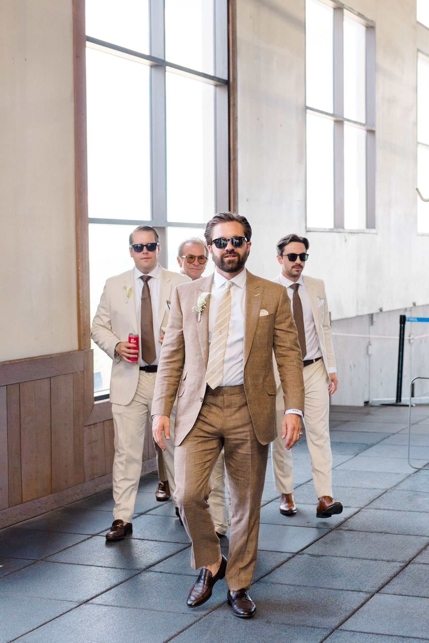 Four men in tan suits and sunglasses walk confidently through a bright indoor venue, led by a groom in a brown suit.