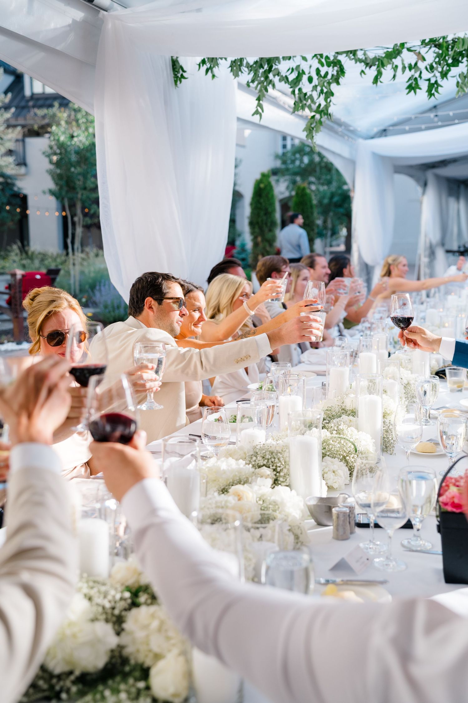 Guests raising glasses in a toast at an elegant outdoor wedding reception with white floral centerpieces.