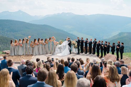 Outdoor mountain wedding ceremony with bridal party in gray dresses and black suits against scenic Colorado peaks.