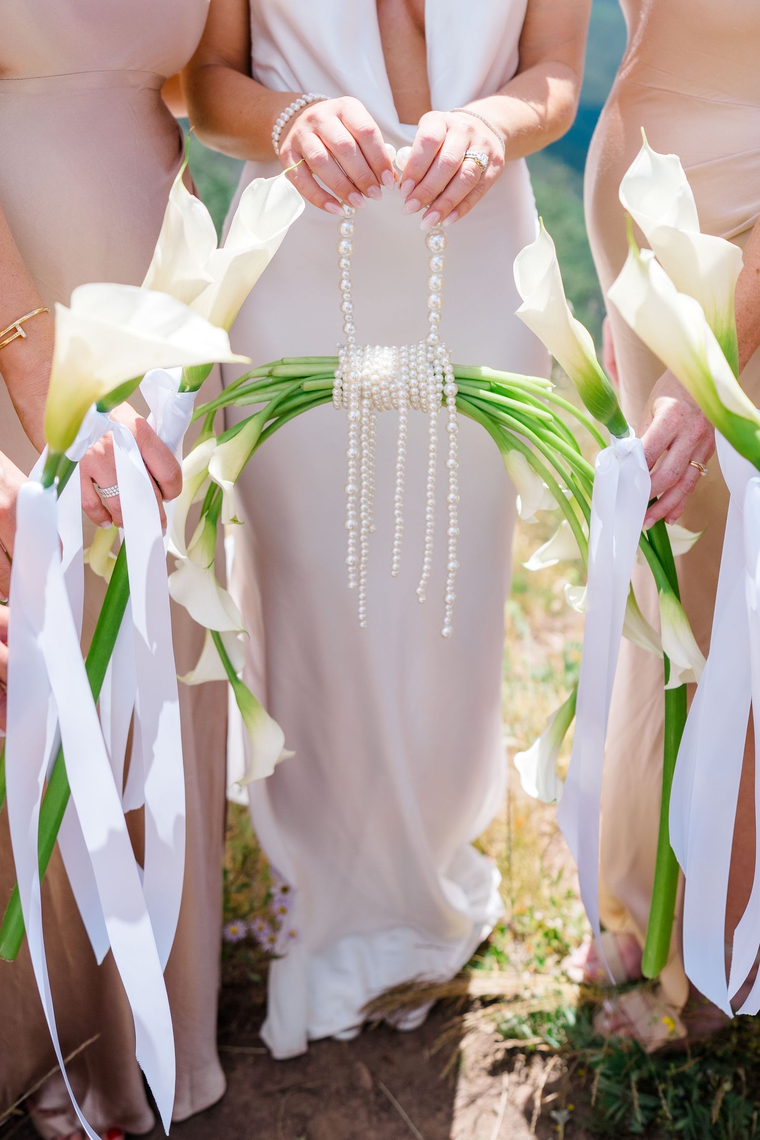 Bridesmaids holding elegant white calla lily bouquets with pearl accents and white ribbons at a wedding ceremony.