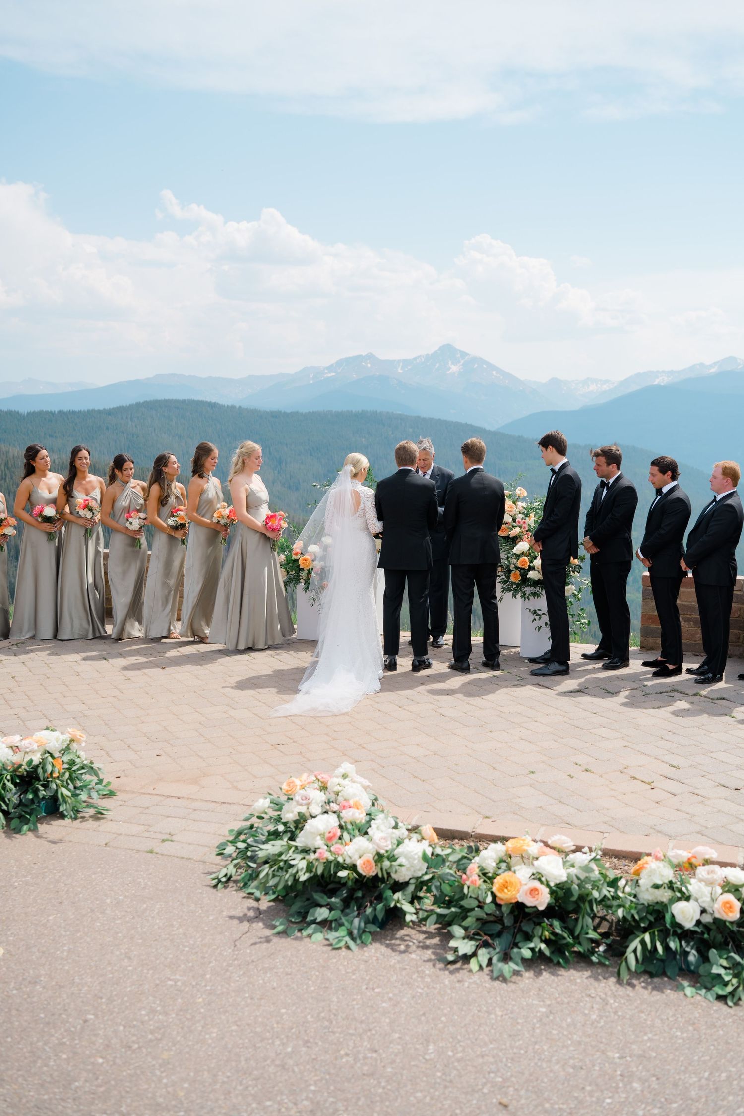 At Vail Wedding Deck outdoor mountain wedding ceremony with bridesmaids in gray, groomsmen in black, and floral arrangements under sky.