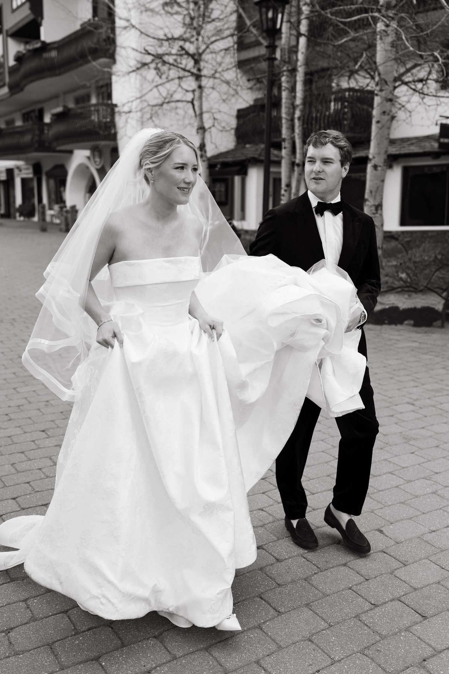 Black and white photo of a smiling bride in a white gown and veil, with a groom in a tuxedo carrying her dress train.