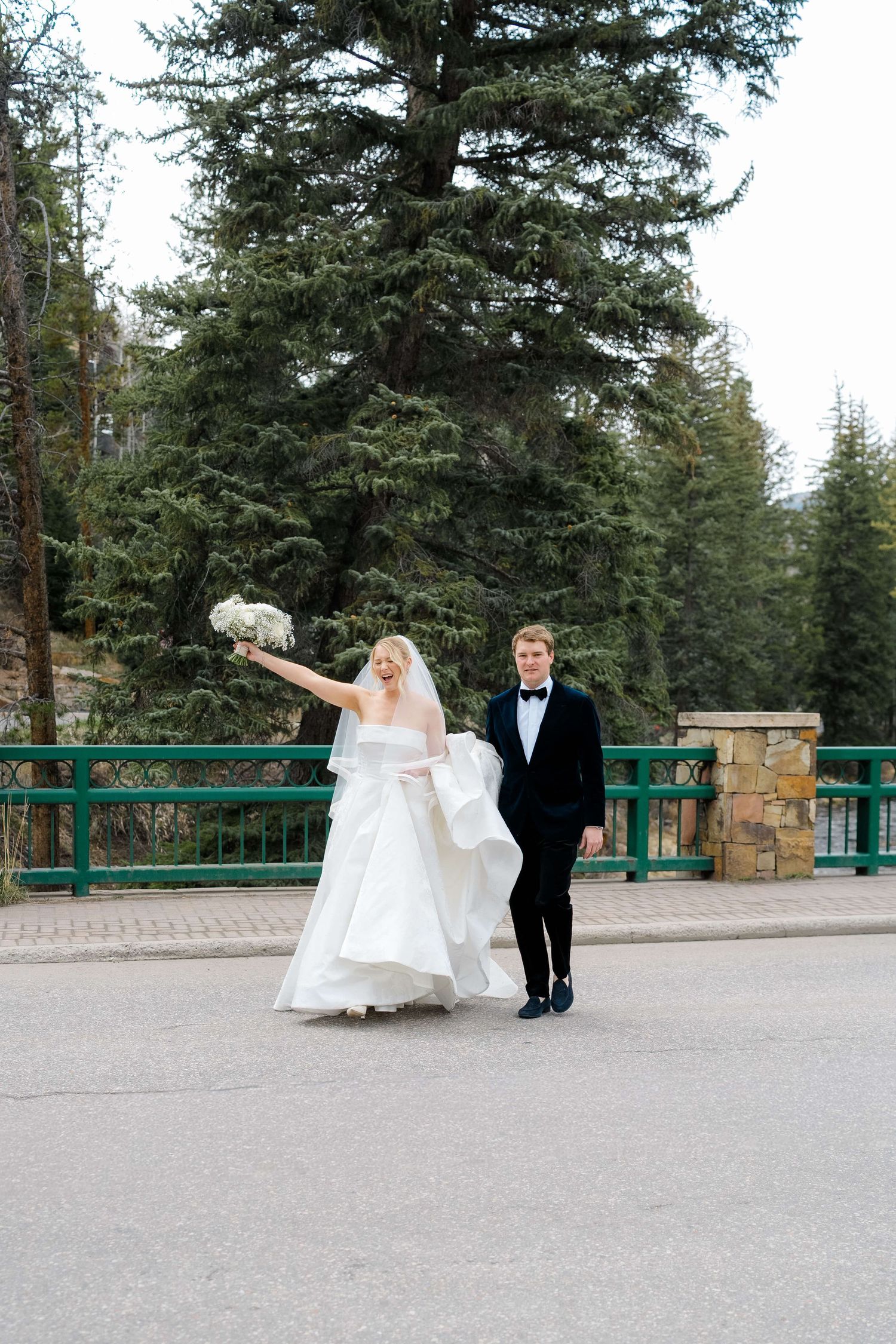 Bride and groom walking joyfully on a bridge surrounded by tall pine trees, bride raising bouquet in celebration.