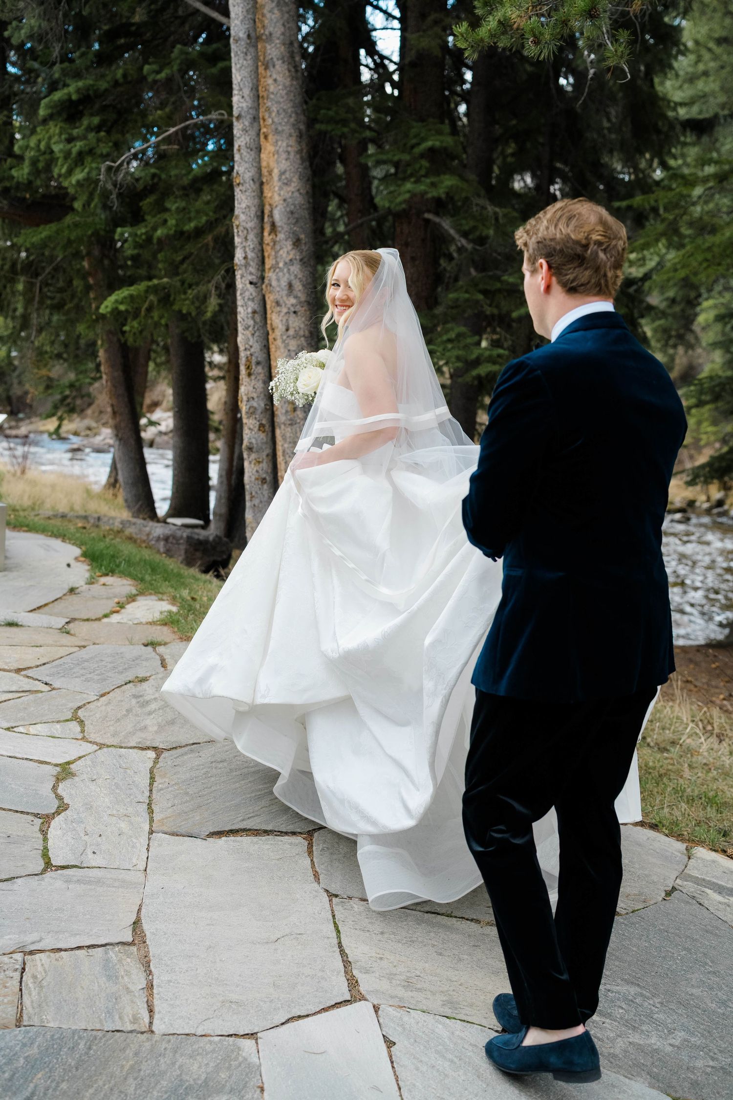 Bride in flowing white gown and veil walks with groom in navy suit on stone path among pine trees at Vail Sonnenalp Wedding.